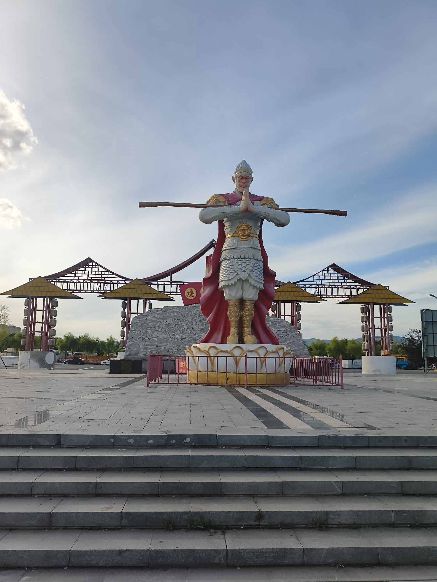 The image depicts a large statue of Sun Wukong, the Monkey King, from the Chinese classic novel Journey to the West. He is depicted in a traditional warrior's stance, with his hands clasped in prayer and a golden staff held across his shoulders. The statue is colorful, with a red cape, golden armor, and white fur details on his face. It stands on a tiered, lotus-shaped pedestal within an open, paved plaza.
Behind the statue, there are several large, pagoda-like structures with yellow roofs and red supporting beams. A stone-like structure, possibly a backdrop or part of the scenery, is visible behind the pedestal. Red fencing surrounds the immediate area of the statue. The plaza is paved with large, gray stone tiles. In the foreground, there are wide stone steps leading up to the plaza level.
The environment is outdoors, under a partly cloudy sky with blue tones. In the background, there are trees and distant hills. Some vehicles are visible in the mid-ground, suggesting the plaza is located in an area with road access. The overall impression is of a public monument or park area dedicated to the character of Sun Wukong.
The location is identified as 新城村 (Xincheng Village), which translates to "new city village" or "new town village." Given the context of the statue and its setting, this likely refers to a park, scenic spot, or cultural attraction within or near this village.