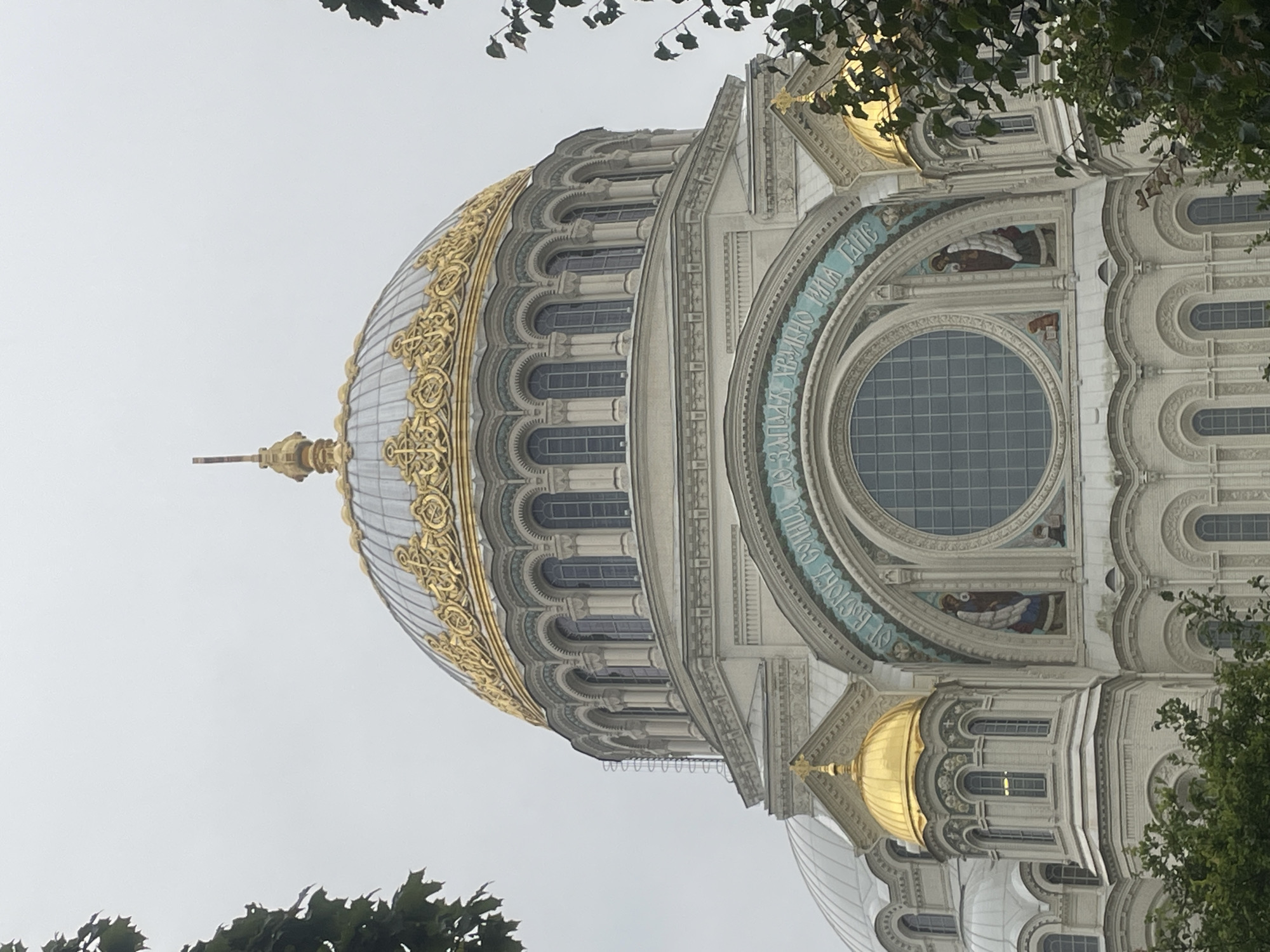 The image shows the Naval Cathedral of Saint Nicholas in Kronstadt, Russia, under an overcast sky. The grand cathedral features a large, ornate golden dome with intricate decorative patterns and a cross at its apex. Below the main dome are multiple tiers of arched windows supported by columns. Surrounding the central dome are smaller golden domes and white architectural elements adorned with decorative carvings. A large circular window is visible on the facade, framed by a mosaic inscription in Cyrillic script and depictions of figures. Green foliage from trees partially obscures the lower portions of the building on the left and right sides of the image. The overall scene is a detailed view of the cathedral's exterior architecture.