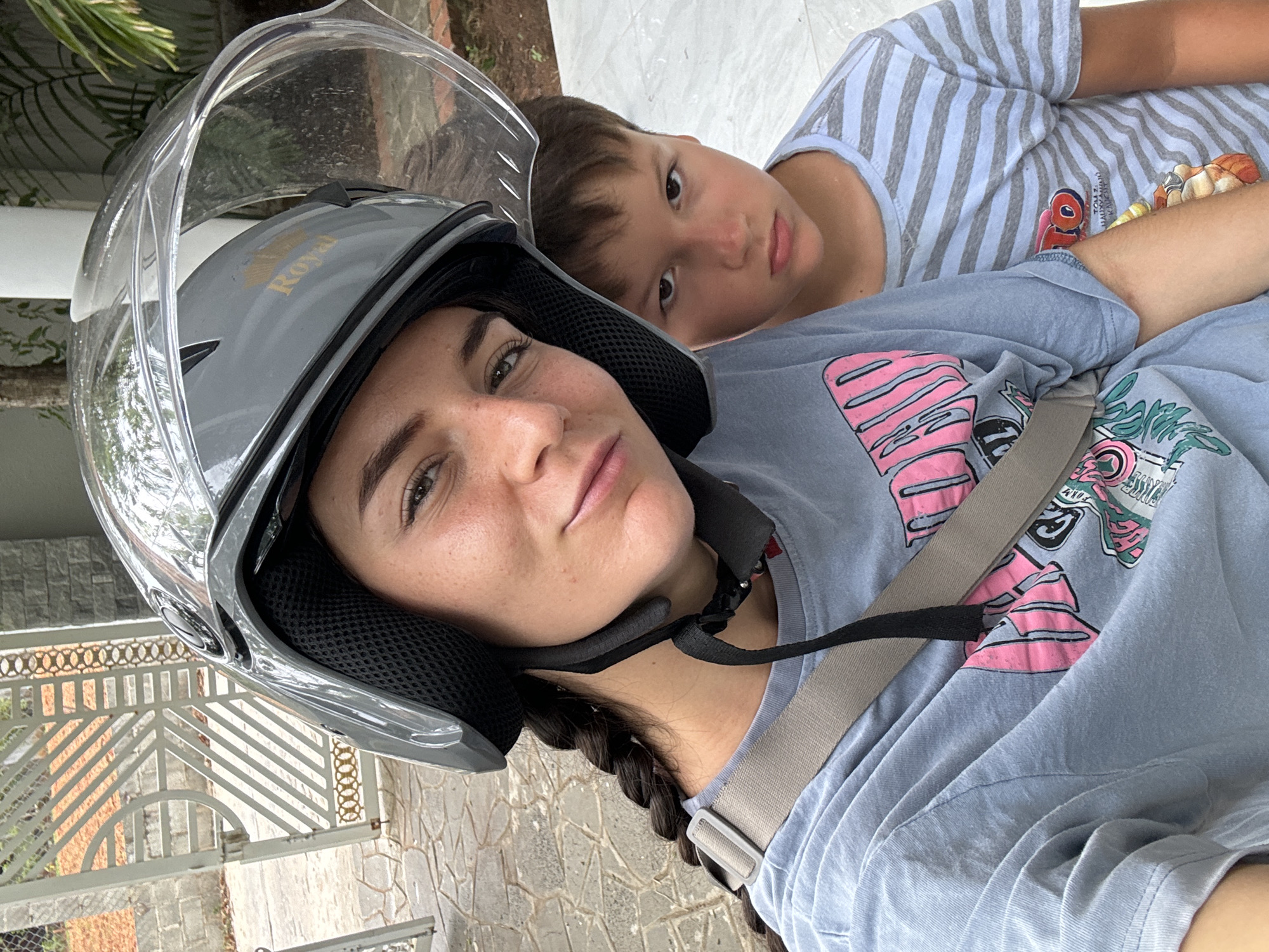 A medium shot depicts a young woman and child in Mũi Né, Vietnam. The woman, angled slightly towards the viewer, wears a gray full-face motorcycle helmet with a clear visor. Her dark hair is braided and pulled to the side. She is wearing a blue T-shirt with a graphic design. A seatbelt is visible across her body. Beside her, a young boy with brown hair and eyes looks towards the camera. He is wearing a striped blue and white T-shirt. The background includes a fence and foliage. The location is outdoors. The perspective is slightly angled, as if the photograph was taken from a higher position.