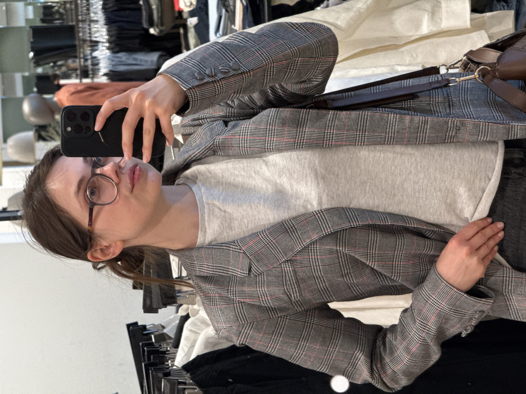 A young individual is captured taking a mirror selfie in what appears to be a clothing store or fitting room located in Budapest, Hungary. The person, appearing female, holds a black smartphone with a multi-lens camera system in their right hand, pointing it towards the mirror to capture their reflection. They are dressed in a grey plaid blazer featuring subtle red and white lines, worn over a light grey t-shirt. A dark brown handbag with a strap is visible over their left shoulder. The individual wears round, thin-rimmed glasses and has light brown hair tied back, with a small ponytail visible from the side. In the background, clothes racks filled with various garments and folded clothes are visible, confirming the retail setting. The lighting is bright and even, characteristic of an indoor commercial space. The person's expression is neutral, focused on the act of taking the photograph. No discernible text is visible on their attire or the phone screen.