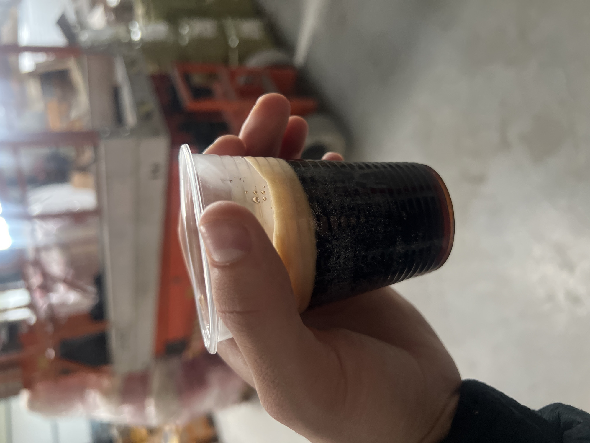 A hand holds a clear plastic cup containing a dark-colored beverage with a tan layer on top. The background is blurry and appears to be an indoor space with shelving and equipment. The exact location in Burgas, Bulgaria cannot be determined from the image.