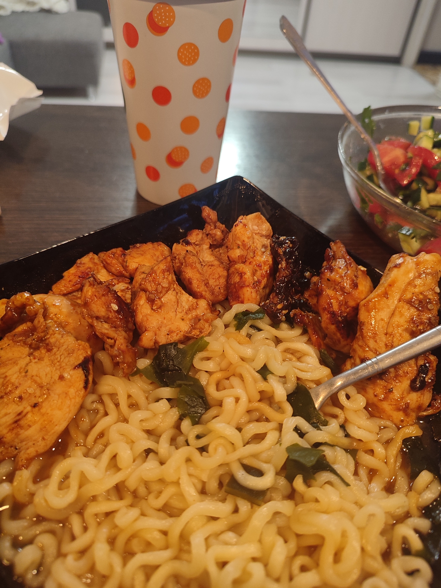 A close-up shot captures a black plate filled with ramen noodles and chicken. The noodles are a pale yellow and coiled, with some green vegetable matter mixed in. Several pieces of cooked chicken, glazed and slightly charred, are arranged on top of the noodles. To the right of the plate, a glass bowl contains a salad of chopped tomatoes and cucumbers, with a fork resting in it. In the background, a white cup adorned with colorful polka dots sits on a dark wooden table. The scene is set indoors, likely in a home environment, with a blurred background suggesting a living room or dining area. There are no people visible in the image. The lighting is warm and natural, indicating it might be daytime. There is no information in the image that would allow for the determination of the specific location within Vsevolozhsk, Russia.