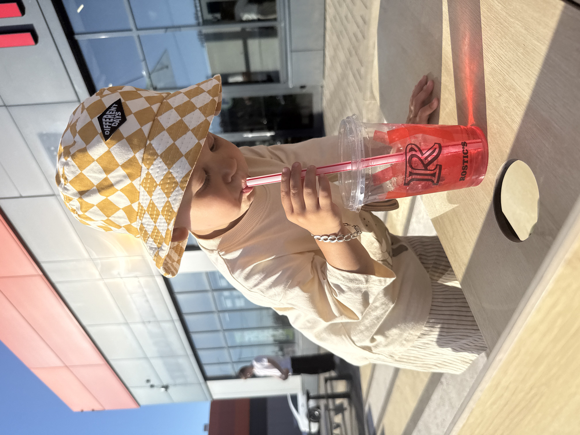 A young child, wearing a checkered bucket hat and a light-colored t-shirt, is shown drinking from a red beverage in a clear plastic cup with a red straw. The cup has a logo that appears to be an 'R' with the word "ROSTIC'S" below it. The child is seated at a light-colored wooden table outdoors, with sunlight casting shadows. In the background, there is a modern building with large windows, and a person in a white t-shirt and dark pants can be seen standing near a white car. The overall setting suggests an outdoor dining area or a public space associated with a restaurant. The image is taken in Шебунино, Russia.