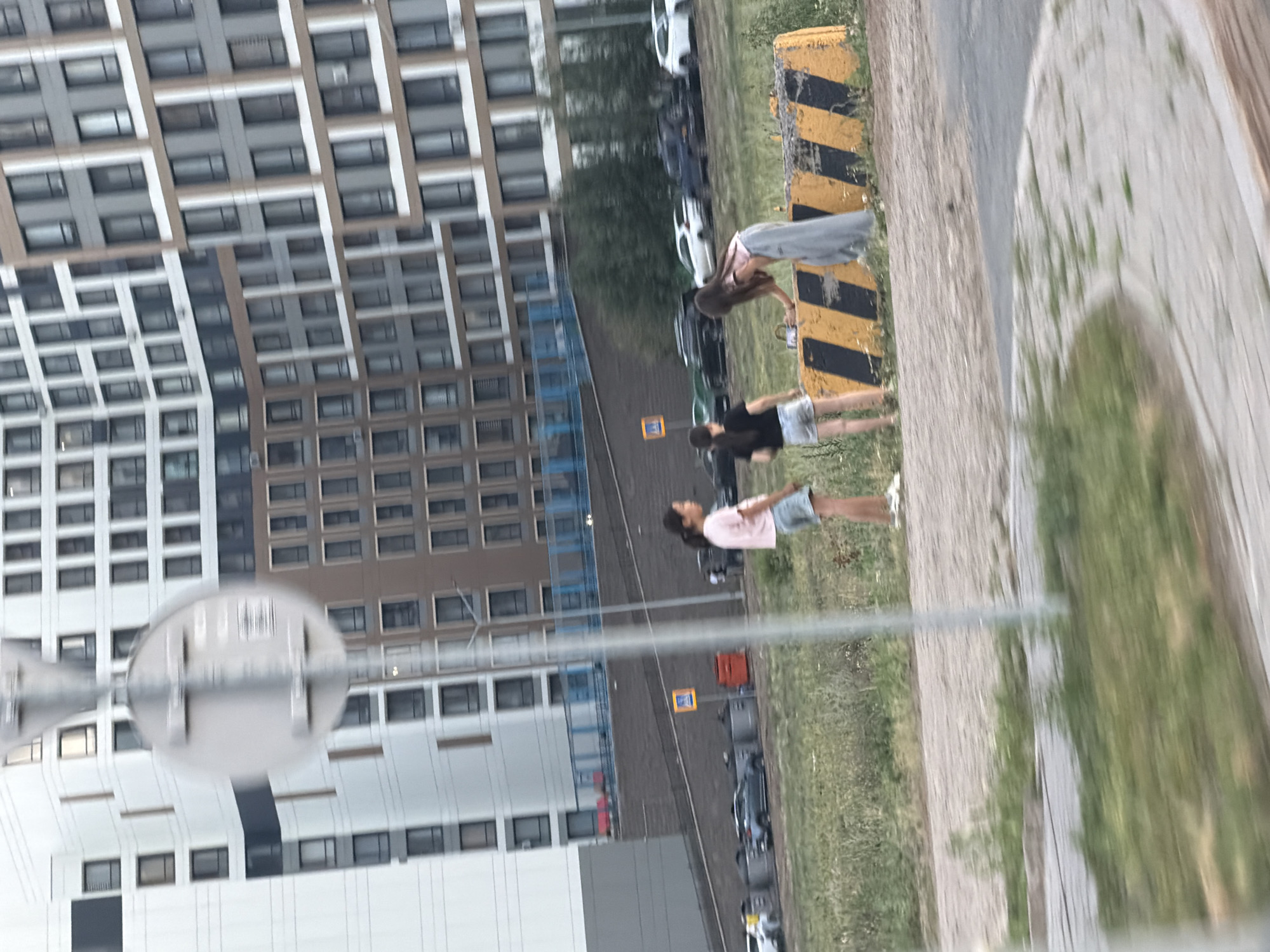 A group of four young people are in an outdoor scene, possibly a park or a street adjacent to a large residential building complex. The building appears to be modern with many windows. The weather is overcast, and the time of day seems to be late afternoon or early evening.
In the foreground, a person wearing a light pink top and denim shorts is looking up and to the right, their body angled towards the right side of the frame. To their left, another person in a black t-shirt and denim shorts is visible from the waist down, also facing right. Further to the right, a person in a light grey skirt and a pink top is bending down, reaching towards something on a yellow and black striped barrier. This barrier seems to be a road divider or a safety feature. Another person, wearing a light grey top and a light blue skirt, is standing further back, also facing right, with their head tilted up.
The ground is a mix of pavement and grass. In the background, several cars are parked or driving on a road. There is a yellow sign with a blue symbol visible on the right side of the frame. The image is rotated 90 degrees clockwise.