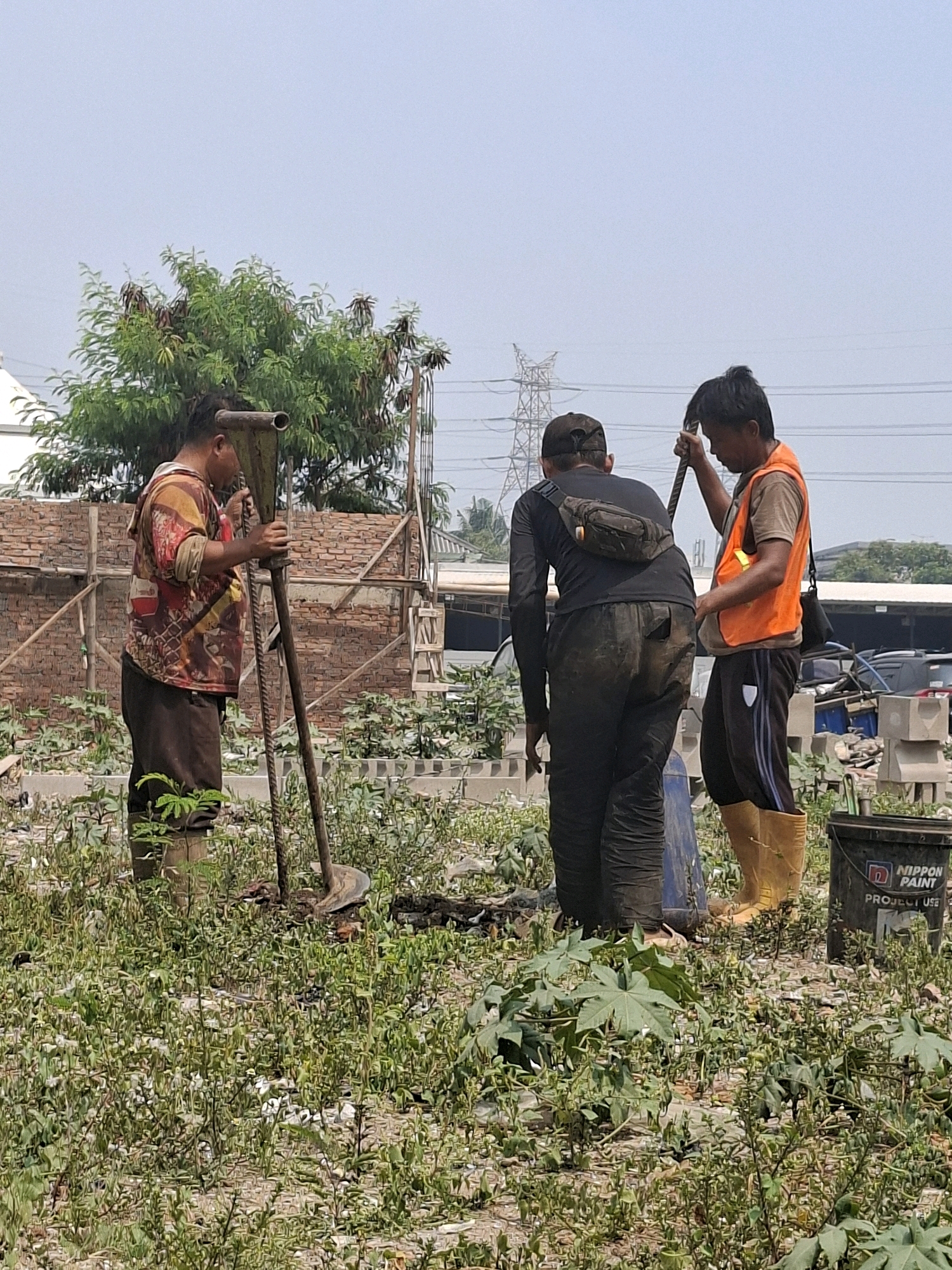 Three men are working in an outdoor construction site in North Jakarta, Indonesia. The foreground is filled with lush green vegetation and rough terrain. In the middle ground, the men are engaged in an activity involving a large, metallic tool that resembles a manual earth auger. One man on the left is operating the tool, dressed in a colorful patterned shirt and brown pants with rubber boots. A second man in the center, wearing a black long-sleeved shirt and black pants, is also positioned near the tool. The third man on the right is wearing an orange safety vest over a dark shirt, dark pants, and yellow rubber boots. He is holding a metal rod. In the background, there is a partially constructed brick wall, wooden scaffolding, a large tree, and in the distance, power lines and a tall transmission tower are visible. The sky is overcast. A black bucket with the "NIPPON PAINT" logo is visible in the lower right corner.