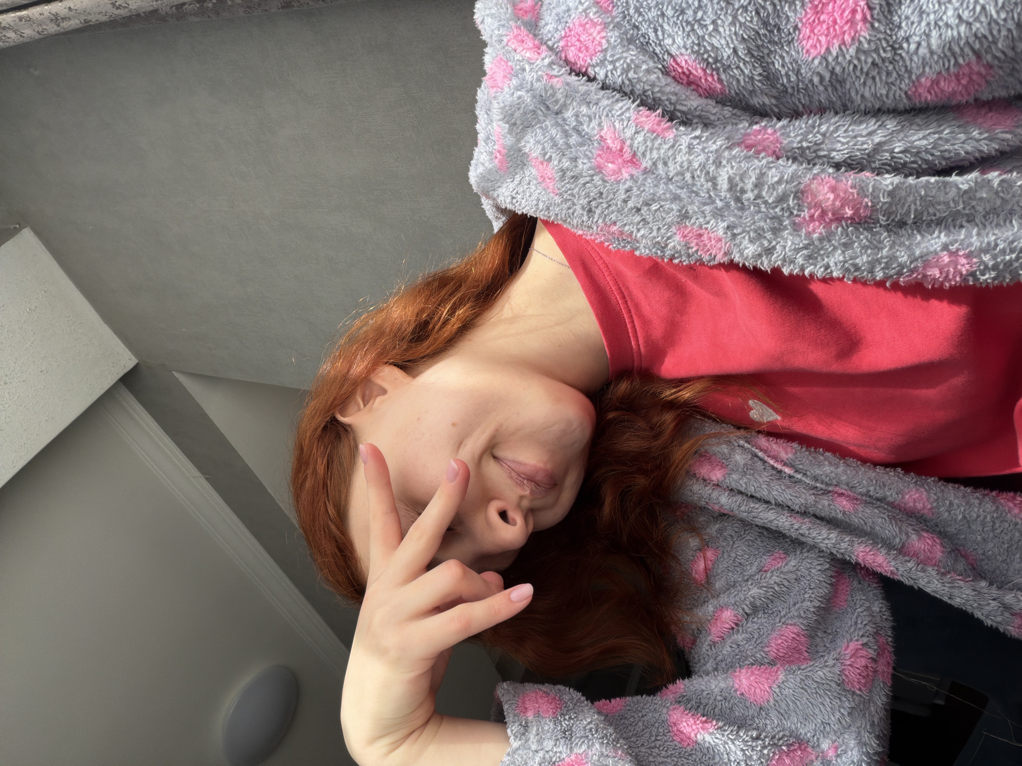 A woman with red hair, wearing a red shirt and a gray robe with pink hearts, is lying down and holding up a peace sign. The background includes a gray ceiling and a white wall with a light fixture. I have no information about this photo being taken in Tbilisi, Georgia.