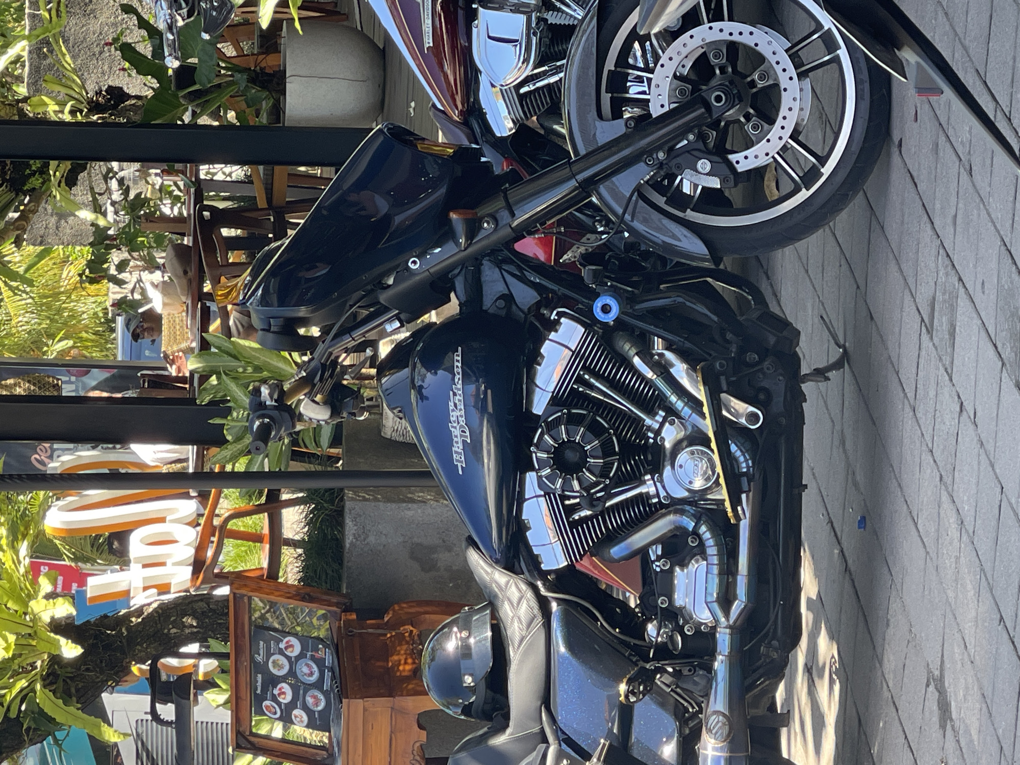 The image shows a dark-colored Harley Davidson motorcycle parked on a stone-paved surface, likely a sidewalk. The motorcycle is angled diagonally across the frame, with the front wheel and engine prominently displayed. The motorcycle has a black windscreen. Part of another motorcycle is visible behind it. The environment suggests an urban setting, with a sign on a brown frame and vegetation partially visible in the background. There are plants and a small tree. The lighting suggests daytime.