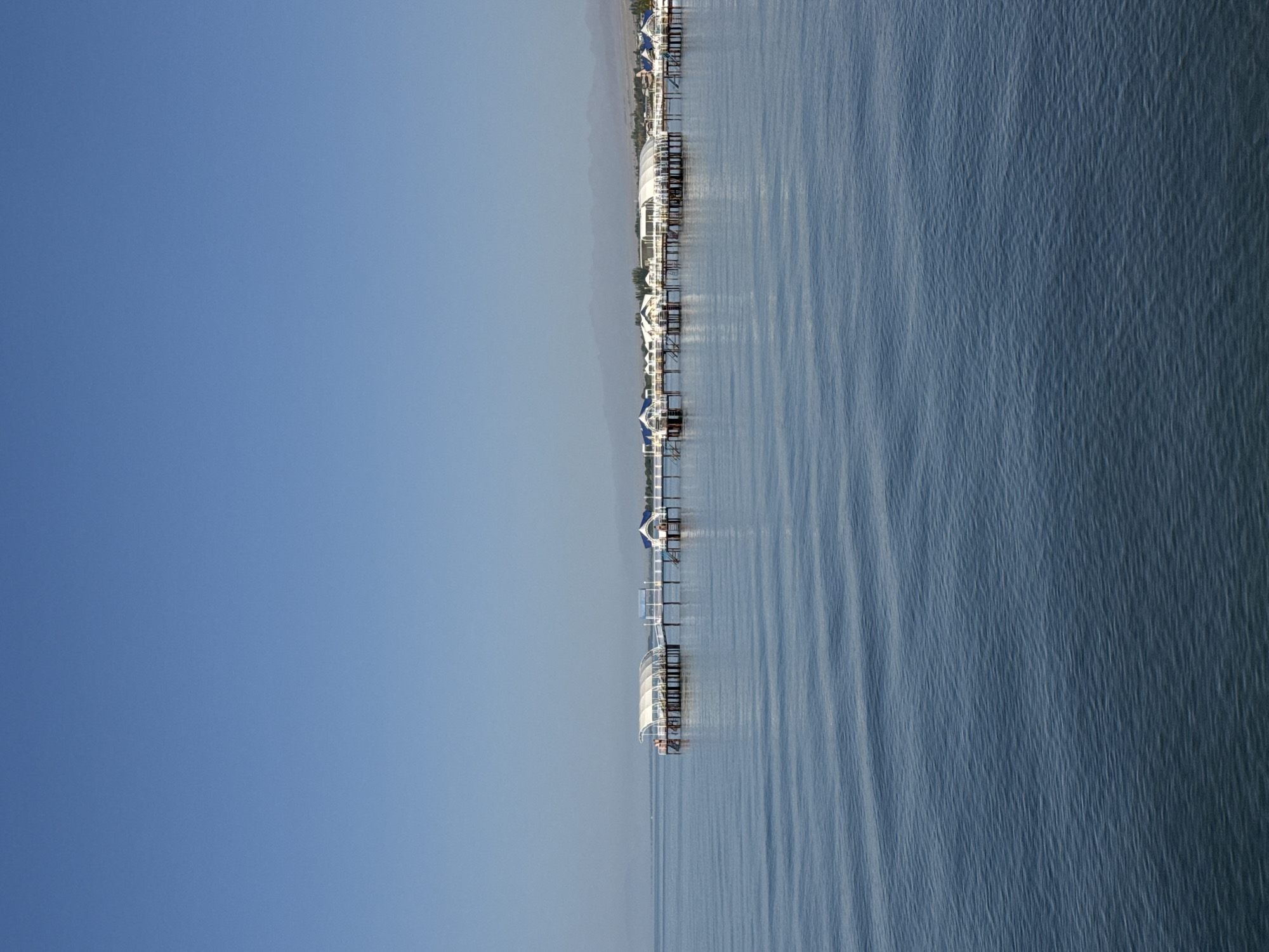 The image depicts a scenic view of a lake with a pier extending into the water, featuring several overwater structures. The structures appear to be small buildings or cabins with white roofs and white railings, supported by stilts. Some of these structures have covered walkways connecting them. The water is calm with gentle ripples, reflecting the clear blue sky. In the distance, a hazy mountain range is visible along the horizon, with a strip of land and trees situated between the lake and the mountains. The location is identified as the city of Sary-Oy, Kyrgyzstan. There are a few individuals visible on the pier, seemingly engaged in casual activity. The overall atmosphere is serene and peaceful, characteristic of a lakeside resort or recreational area.