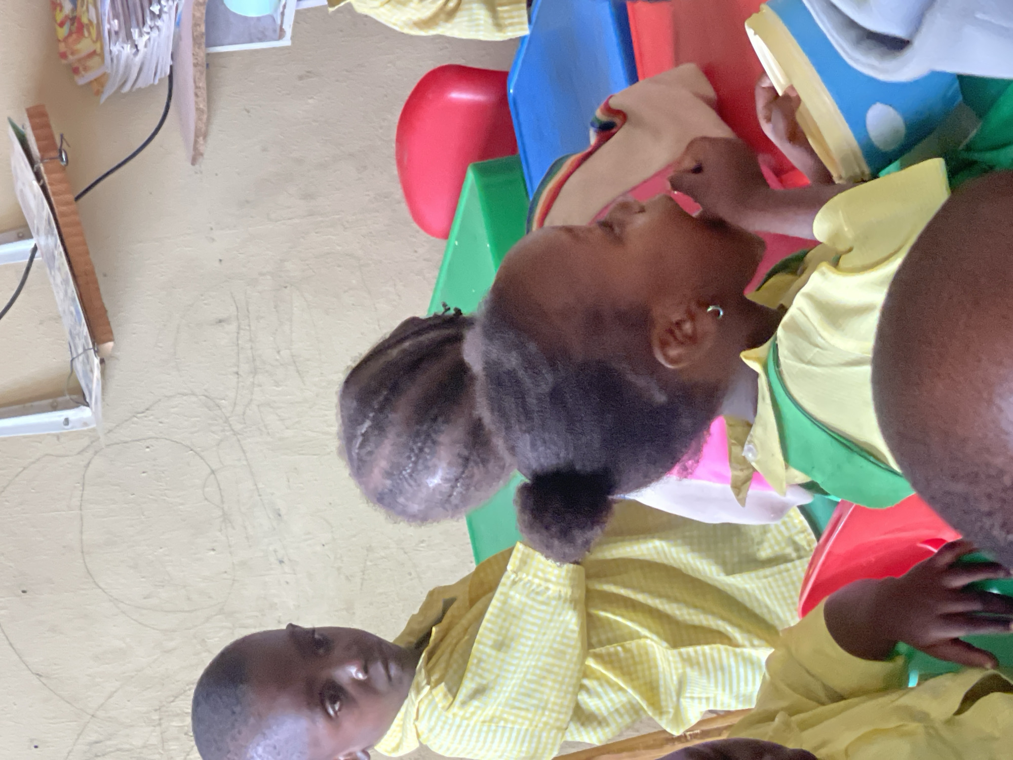 The image depicts a scene in Akwanga, Nigeria, where several children are gathered indoors. The children, dressed in yellow and white checkered uniforms, are seated in an arrangement of colorful plastic chairs in red, blue and green. One child is holding a blue and white book and appears to be reading. The indoor environment has light yellow walls. There are some papers clipped to the wall, with a wire or cord running down the wall.