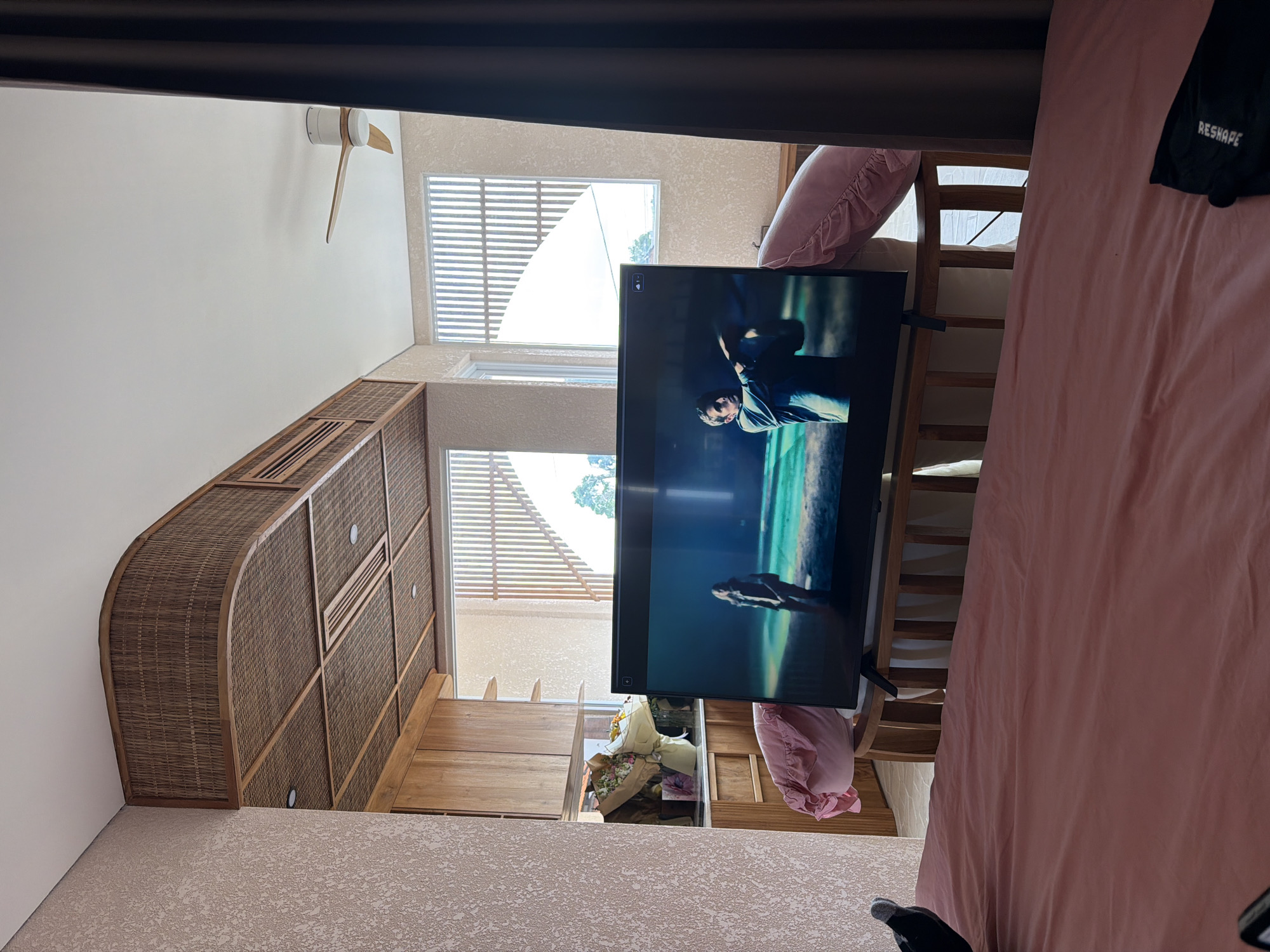 The image depicts a bedroom interior in what appears to be a building in Mengwi, Indonesia. The room features a bed with pink linens and pillows, positioned next to a wooden platform structure. A flat-screen TV is mounted in front of a window, displaying an image of a person. Natural light filters into the room. The room features custom-designed furniture with wicker and wood accents. The overall setting suggests a modern, well-designed bedroom space in Mengwi.