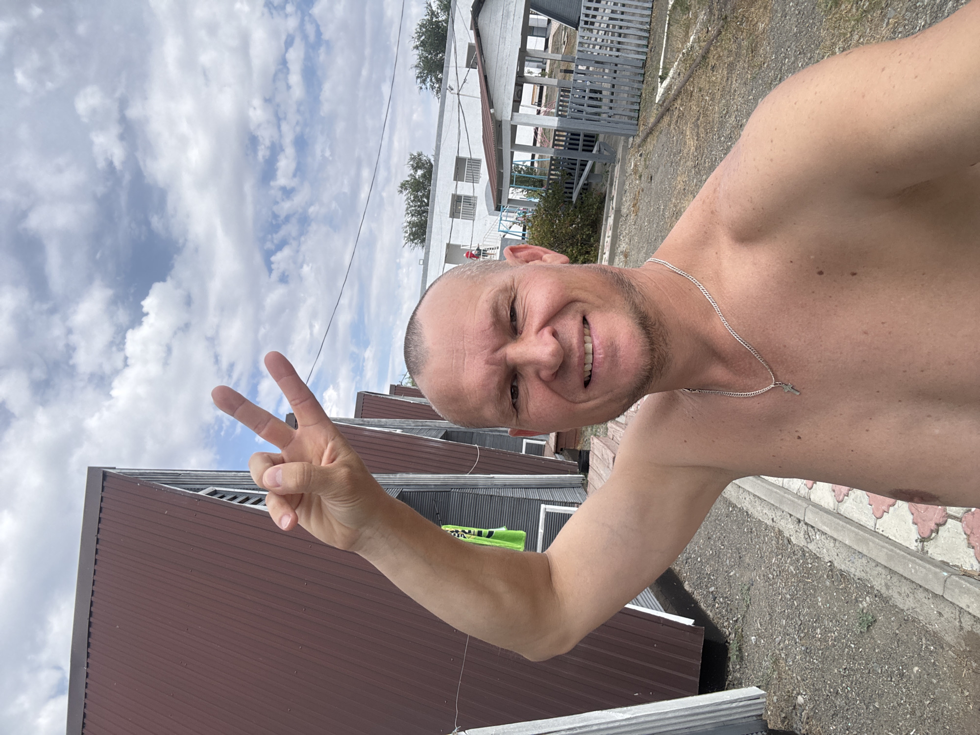 A bald, shirtless man with a light tan smiles at the camera, holding up his right hand in a peace sign. He is wearing a silver chain with a cross pendant. The man appears to be outdoors, with a cloudy sky visible above. In the background, there are several small, brown, A-frame structures with metal siding, possibly cabins or sheds, lined up along a gravel path. Further in the background, a white building with windows and a smaller, white, open-sided structure with a grey fence can be seen. The ground is covered in gravel and some patchy dry grass. The scene appears to be in a rural or semi-developed area.