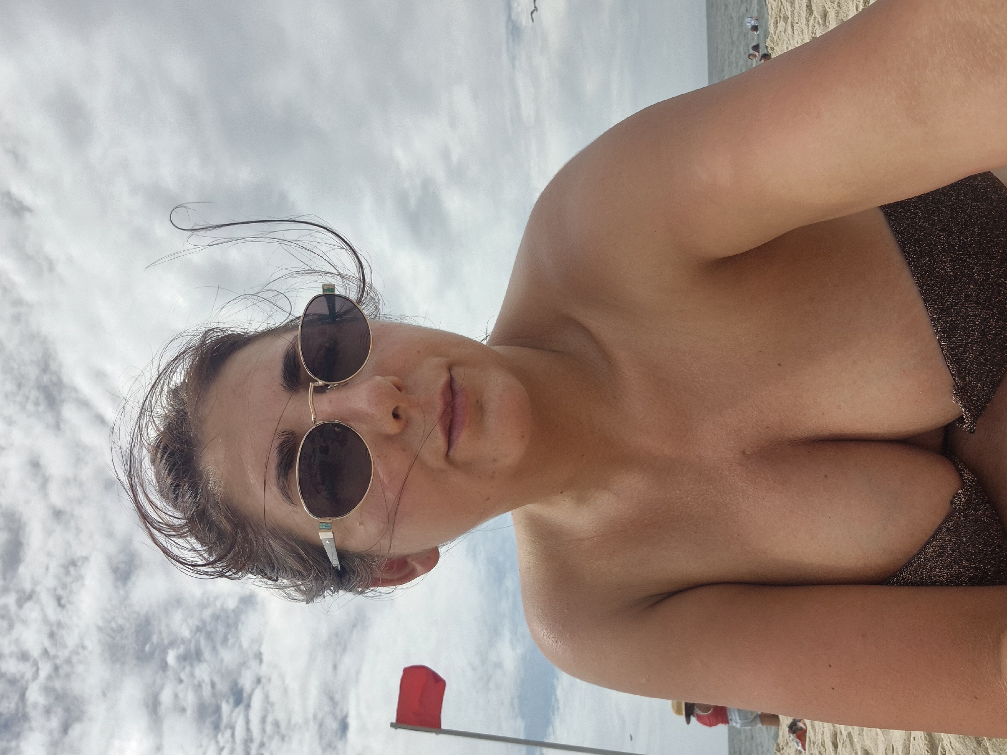 An overhead selfie taken by a woman on a cloudy day at the beach. The woman is wearing sunglasses and a dark bikini top. She has brown hair that is blowing in the wind. In the background, a red flag is flying on a pole, and there are people in the distance swimming in the ocean. The sand is visible in the foreground. The image appears to have been taken at Sandy Hook, a section of Gateway National Recreation Area in Middletown Township, New Jersey.