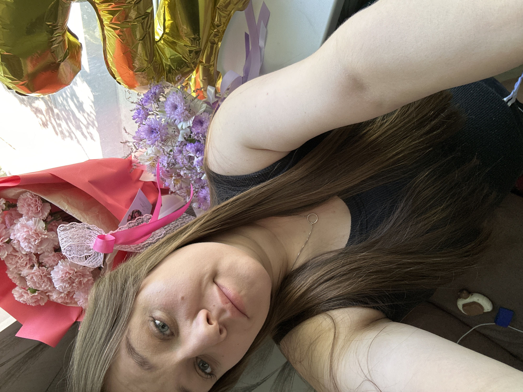 The image shows a selfie of a woman with brown hair, wearing a black top and a silver necklace. She is positioned at an angle, with her face tilted towards the camera. She is surrounded by several objects. To her left, there are two bouquets of flowers; one is composed of pink carnations wrapped in pink paper and the other has purple and white flowers. Beside the flowers are two golden number balloons, "20". In the background to her right, partially visible, is a small white and brown object and a blue object on a dark brown surface. It is impossible to verify that this image was taken in the city of Chaikovsky, Russia.