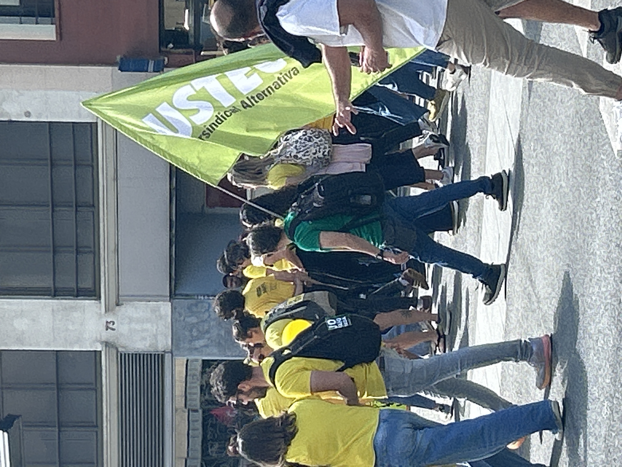 A group of people are marching in a street protest. They are holding a green flag with the words "USTEC·sindicat Alternativa" written on it. Most of the protesters are wearing yellow t-shirts. The setting is an urban environment, specifically in Barcelona, Spain, as evidenced by the building's architecture and the "73" number plate.