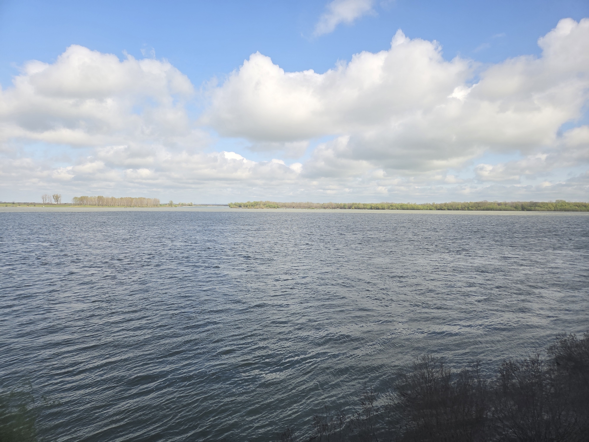 The image shows a wide expanse of water under a partly cloudy sky. The water is choppy with small waves, and its color ranges from gray to blue. In the distance, across the water, there is a low-lying landmass with trees. The sky is blue with numerous white and gray clouds scattered across it. The bottom right corner of the image shows some dark, indistinct vegetation. The location is Somovit, Bulgaria, based on the coordinates.