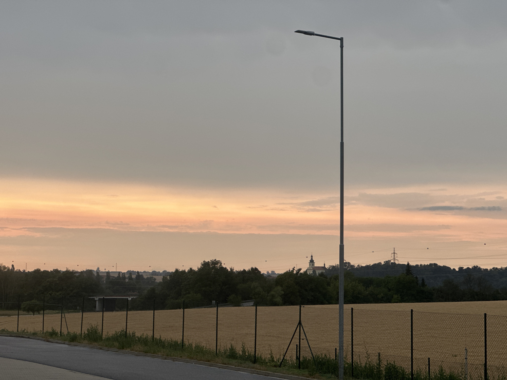 This image depicts a rural landscape at dusk in Brodce, Czechia. The foreground features a paved road with a black wire fence running along its edge. Behind the fence, a field of dry, golden vegetation, likely harvested crops, stretches into the distance. The middle ground is characterized by a dense line of green trees, with a small white structure, possibly a building or a bridge, partially visible amongst them on the left. Further back, a distinctive church steeple with a cross can be seen rising above the treeline, suggesting a village or small town. The sky is a dramatic canvas of soft grays and muted oranges, indicating the time of day as sunset or twilight. The clouds are present but appear diffuse, contributing to a slightly overcast or hazy atmosphere. A tall, solitary lamppost stands prominently in the right half of the frame, its dark silhouette stark against the lighter sky. Power lines and a distant transmission tower are visible against the horizon, hinting at the proximity of infrastructure. There are no people or animals visible in the scene. The overall mood is serene and tranquil, characteristic of the quiet transition from day to night in a rural setting.
