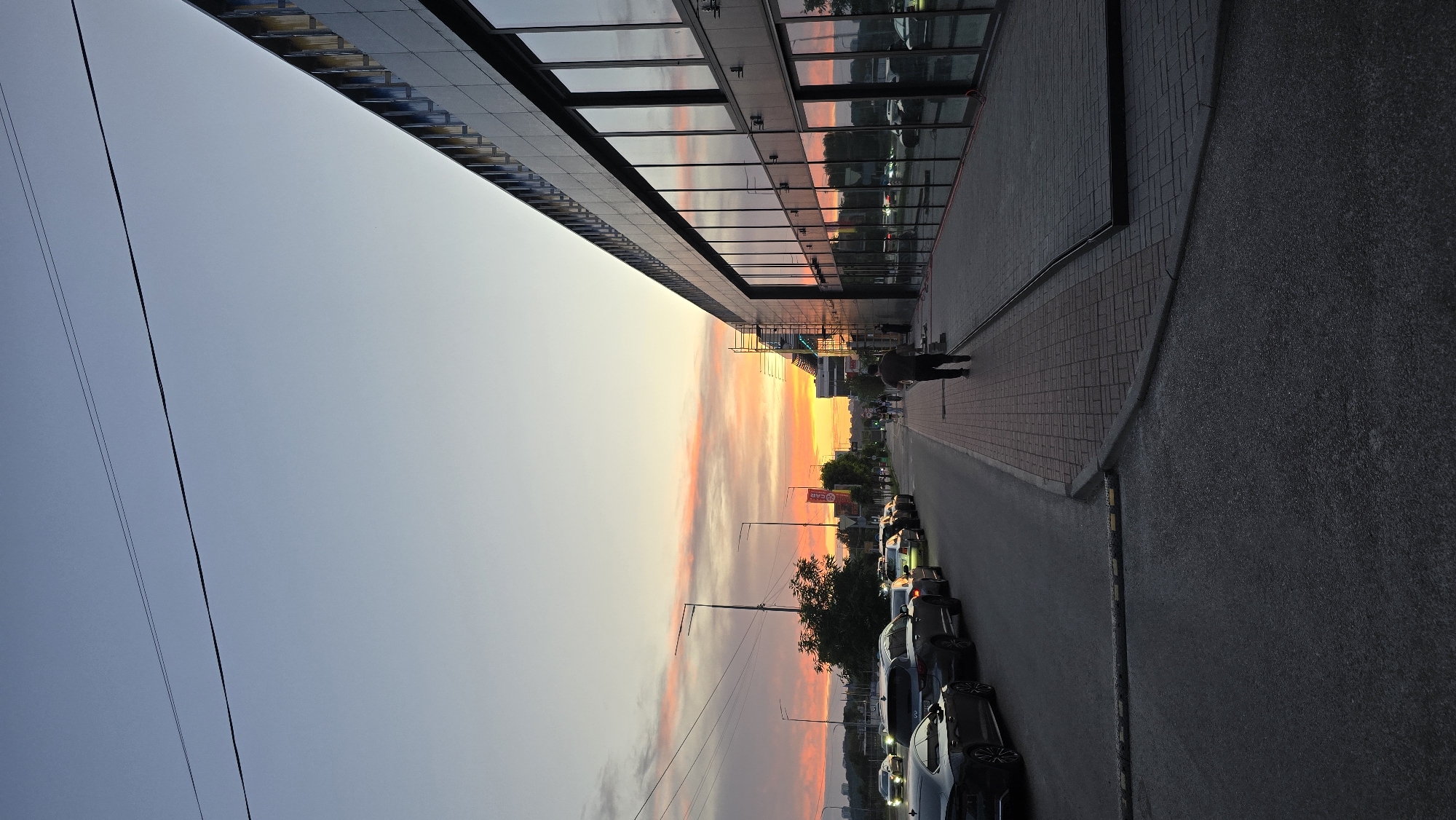 The image depicts a street scene in Kyiv, Ukraine, during sunset. A modern building with a glass facade is on the right side of the frame, reflecting the colorful sky and surrounding environment. A man walks along the sidewalk in front of the building. On the left side of the street, numerous cars are parked in a line, and traffic can be seen in the distance. The sky is filled with dramatic hues of orange, pink, and blue, characteristic of a sunset. Power lines are visible stretching across the upper portion of the image. The street is paved, and a sidewalk made of brick pavers runs alongside the building. The overall atmosphere is urban and serene, capturing the transition from day to evening.