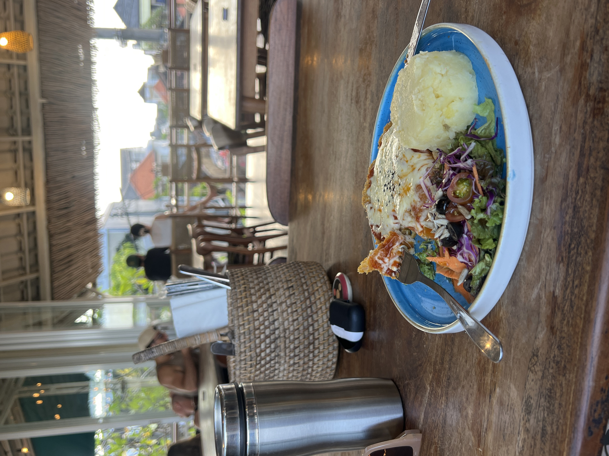 The scene depicts a meal served on a blue-rimmed plate placed on a wooden table, likely in a restaurant in North Kuta, Indonesia. The plate contains mashed potatoes, what appears to be a piece of meat with melted cheese on top, and a salad composed of lettuce, red cabbage, tomatoes, carrots, and olives. A fork is present on the plate, piercing a piece of the meat dish.
Next to the plate is a woven basket containing napkins and utensils. A stainless steel tumbler and a pair of sunglasses are placed on the table near the basket.
Through the glass window behind the table, other tables and chairs are visible, suggesting an outdoor seating area. People can be seen through the window in the background. The outside environment includes buildings and trees.