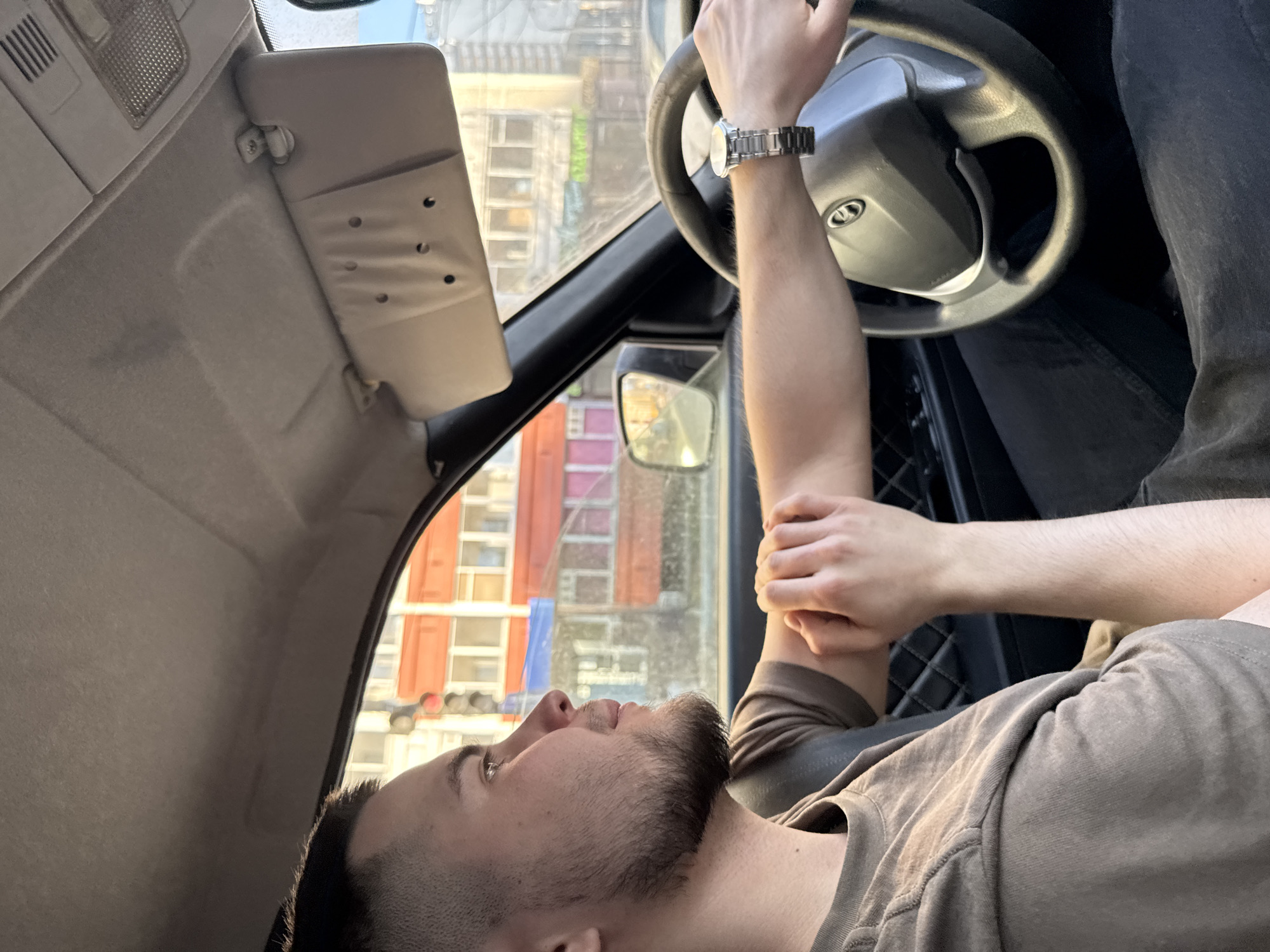 A male individual is observed in profile from the front passenger seat of a left-hand drive vehicle, positioned as the driver. He is an adult with short hair, a faded haircut style, and a beard, wearing a plain, short-sleeved, muted brown t-shirt. A seatbelt is visible across his left shoulder and chest. His left hand, adorned with a metallic wristwatch featuring a round face and link bracelet, is on the steering wheel. His right hand rests on his left forearm. He is looking forward and slightly right, through the windshield.
The vehicle's interior includes a light grey headliner and a beige passenger-side sun visor, which has three circular indentations. Part of the black dashboard and a quilted pattern on the door panel are visible. The steering wheel is dark with a central emblem, consistent with a Lada vehicle.
Outside the vehicle, an urban street scene is visible under bright daylight, suggesting late afternoon due to the warm light. A prominent multi-story building with a red or orange facade and numerous windows is a central feature. A vertical traffic light displaying a red signal is visible to the left of this building. Other buildings and blurred green signage are also present in the background. A side mirror reflects another vehicle. The scene is located in an urban area of Perm, Russia; however, the precise street or intersection is not identifiable from the visual information provided.