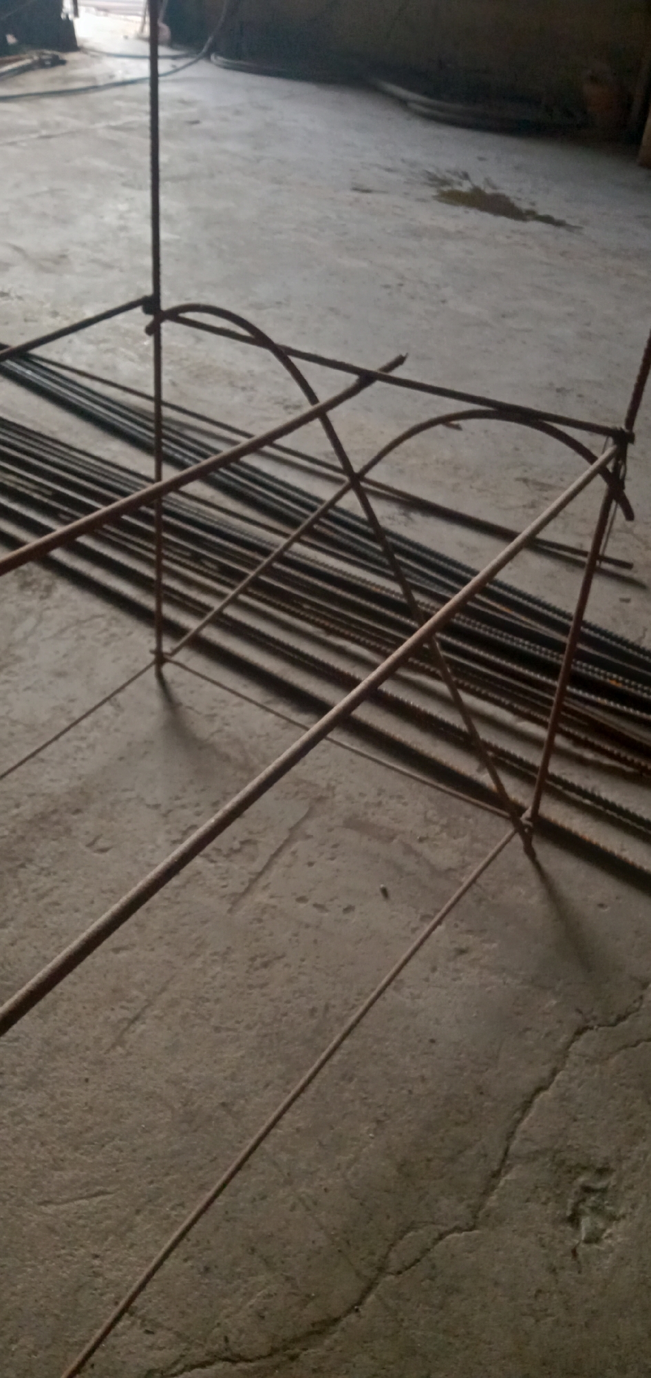 Location: Sumput, Indonesia.
The image shows a set of rusty reinforcing bars (rebar) on a concrete floor. A pre-assembled rebar structure, potentially for concrete reinforcement, stands in the foreground. The structure includes vertical bars and curved horizontal supports. A bundle of straight rebar pieces lies on the floor in the background. There is an area with spilled liquid on the concrete floor. The overall setting suggests a construction site or workshop.