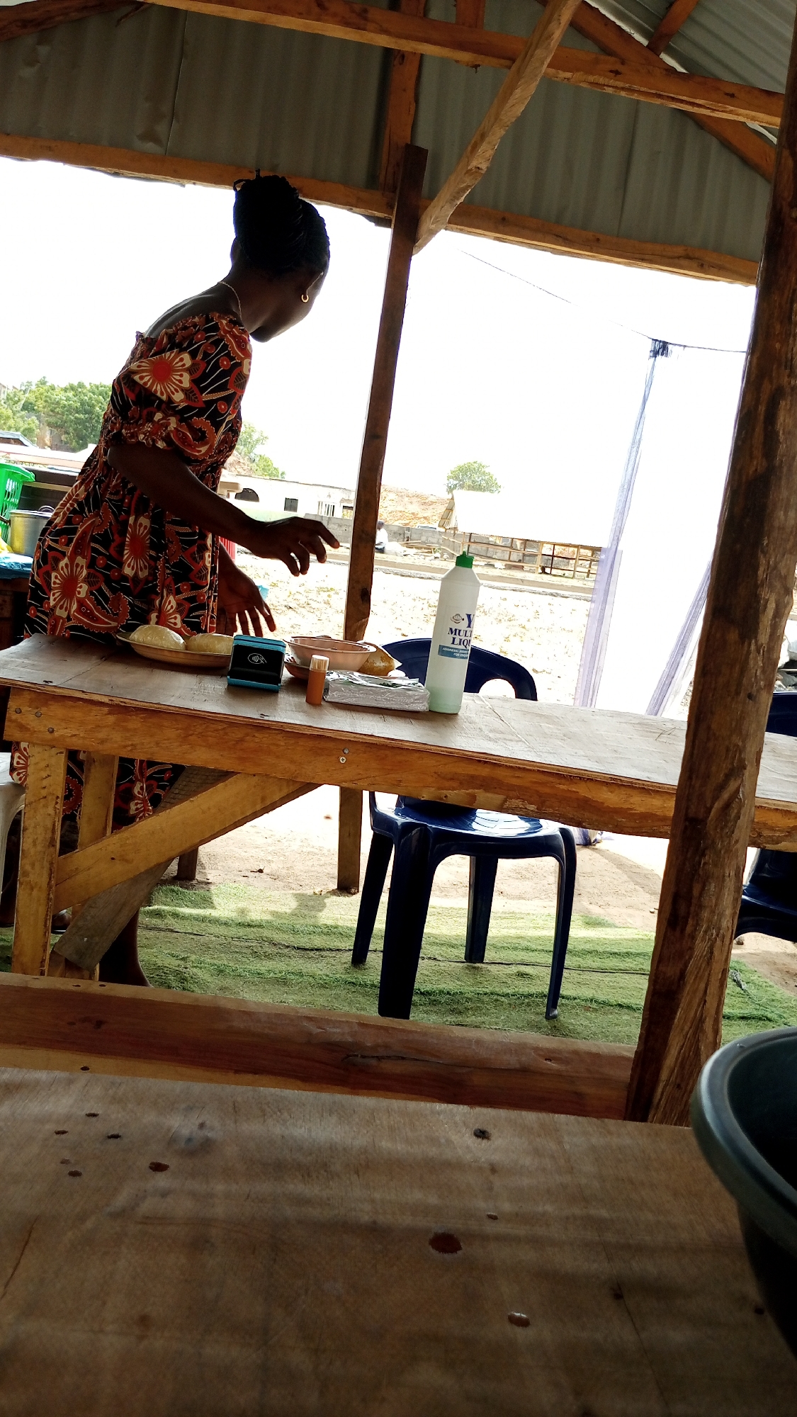 In Lokoja, Nigeria, a woman with an updo hairstyle and wearing a patterned dress stands behind a wooden table. She appears to be preparing food. On the table, there's a plate of what seems to be doughy food, a small electronic device, a jar, and a bottle labeled "MULTI LIQUID." A blue plastic chair is tucked under the table. The scene is under a simple shelter with a tin roof and wooden support beams. In the background, there are buildings and some sparse vegetation. The foreground shows another wooden surface, suggesting this is a simple food stall.