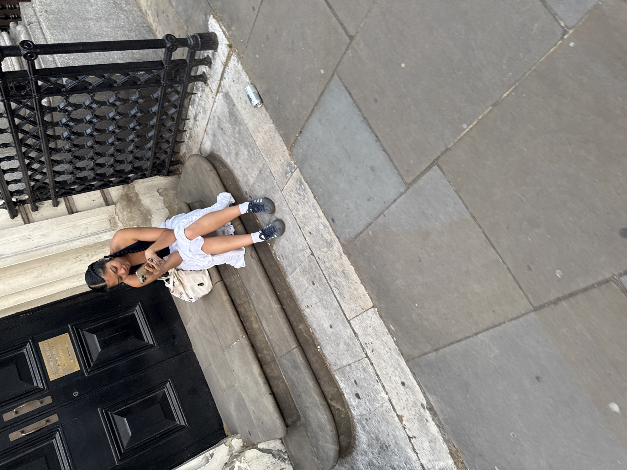 A young woman is sitting on the steps of a building, looking at her phone. She is wearing a black tank top, a white dress, and gray sneakers. A beige backpack is placed on the step next to her. The building has a black door with a brass plaque that has text on it. The steps are made of stone, and the sidewalk is paved with gray tiles. A black metal railing is visible to the left of the woman. The scene appears to be outdoors, likely in a city setting, given the stone steps, sidewalk, and building. The lighting suggests it could be daytime, possibly overcast or a slightly hazy sun. There are no clear indicators of the specific time of day or weather, other than the presence of daylight. The woman's posture and focus on her phone suggest she might be relaxing or waiting.
The text on the brass plaque is partially visible and reads: "THE ROYAL HOSPITAL OF SAINT ELIZABETH OF PORTUGAL AT CHEYNE WALK SW3". The visible text appears to be in English and indicates a historical or institutional building. The context provided states the location is London, United Kingdom, which aligns with the architectural style and the visible text.