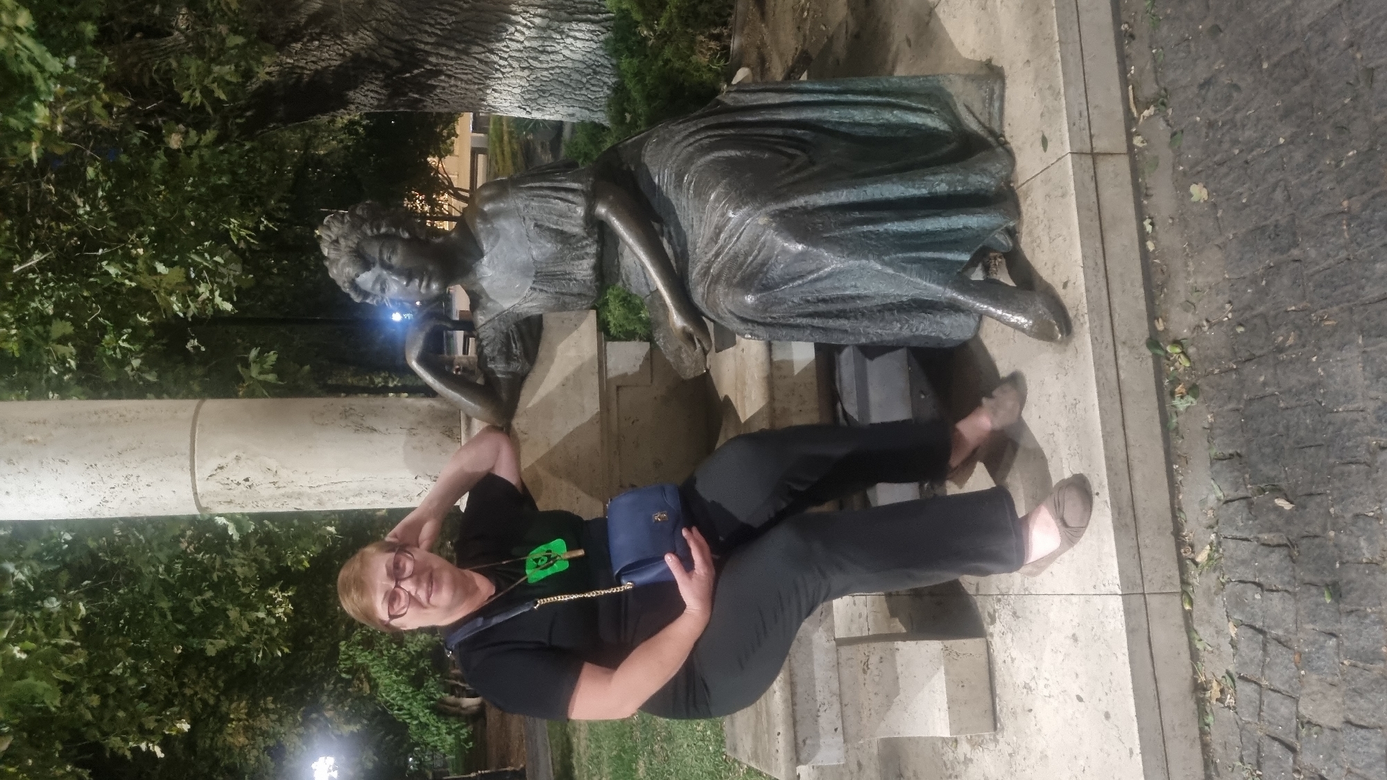 A medium shot captures a woman sitting on a stone bench next to a bronze statue of a woman in a flowing dress. The woman on the left, wearing glasses, a black t-shirt with a green emblem, black pants, and beige flats, rests her head on her hand. She carries a dark blue purse with a gold chain. The bronze statue is positioned to the right, leaning on the bench with one arm, holding a small object in her lap. Both figures are seated on a light-colored stone bench, with a textured cobblestone path visible in the foreground. Lush green trees and foliage form the background, suggesting an outdoor setting, possibly a park or public space. The scene appears to be in Berehove, Ukraine.