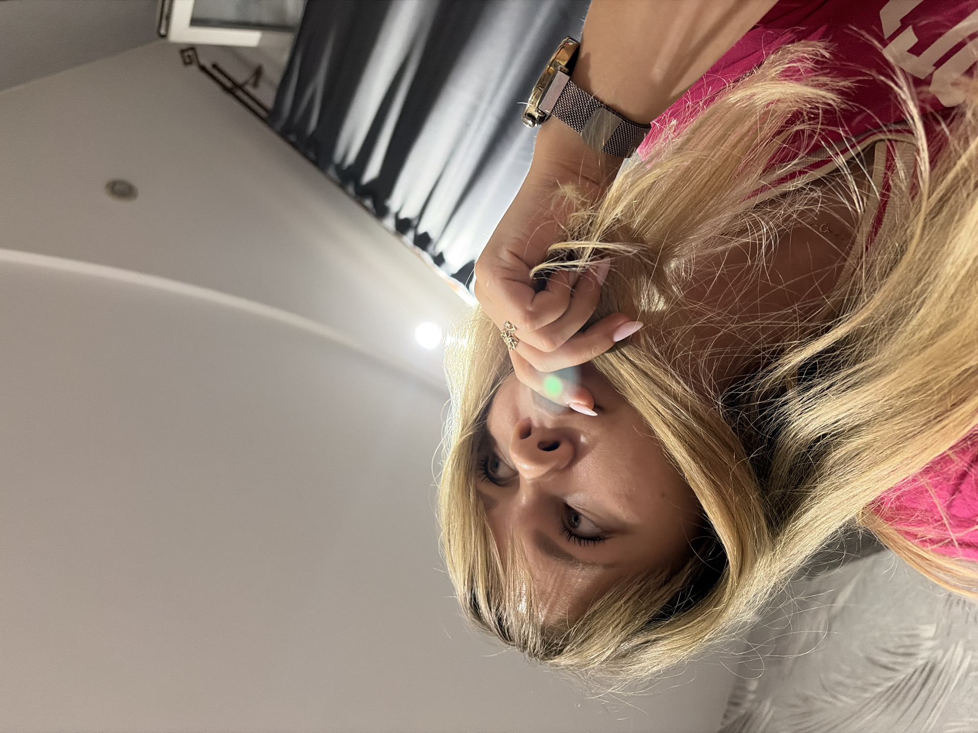 A woman with long blonde hair is facing the camera. She is wearing a pink shirt and a watch on her left wrist. Her left hand is near her face. She has a ring on her finger and appears to have manicured nails. The background shows a gray and white patterned surface, and behind her, there are gray curtains. The location is identified as Chaikovsky, Russia, based on inference from the image and prompt.