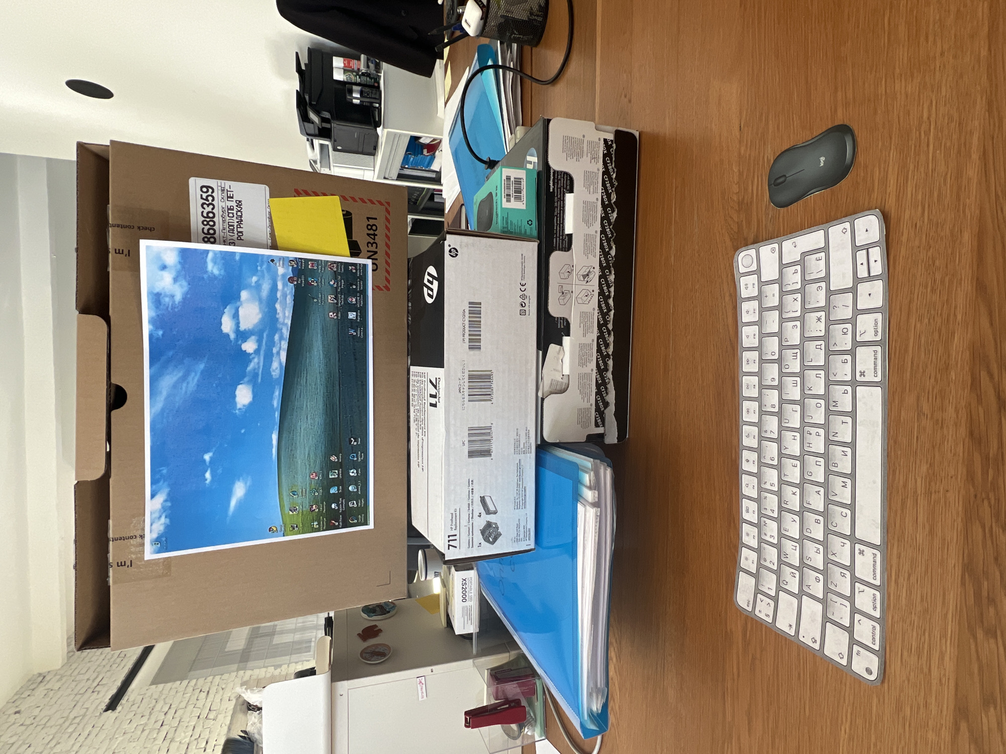 The image displays an indoor desk setting, likely a temporary workstation or a busy home office. The scene is captured with a clear view of a light brown wooden desk surface, illuminated by ambient indoor lighting.
In the foreground, a dark gray Logitech computer mouse and a white keyboard are neatly arranged on the wooden desk. The keyboard features both Latin and Cyrillic characters, including labels like "option," "command," "control," and "fn," indicating its use in a Russian-speaking context.
To the left and center of the frame, a large, open cardboard box is prominent. A shipping label on this box clearly shows Russian text: "нкт-Петербург Склад," "3) (ДОП) СПБ ПЕТ-", "РОГРАДСКАЯ," along with numerical identifiers "8686359" and "N3481," confirming its origin or destination in Saint Petersburg, Russia. Other partially visible text on the box includes "I'm s" and "check content." A yellow sticky note is also attached to this box. Notably, a printed image of a Windows XP desktop background, complete with software icons, is taped onto the cardboard box, playfully serving as a makeshift monitor screen.
Several other boxes and packages are stacked near the large cardboard box. These include HP-branded boxes, with visible product numbers such as "711" and "CF280X," suggesting printer ink or toner cartridges. A smaller turquoise box with a Logitech logo and a white box labeled "XS2000 PORTABLE SSD" are also present.
Further back on the desk, a blue folder or binder filled with papers is visible, next to a red stapler and some decorative items. The background reveals a white wall or cabinet with shelves holding various items, including a black mesh pen holder.
The image portrays a functional workspace, possibly in the process of setting up or organizing computer and office supplies, with strong contextual clues pointing to its location in Saint Petersburg, Russia.