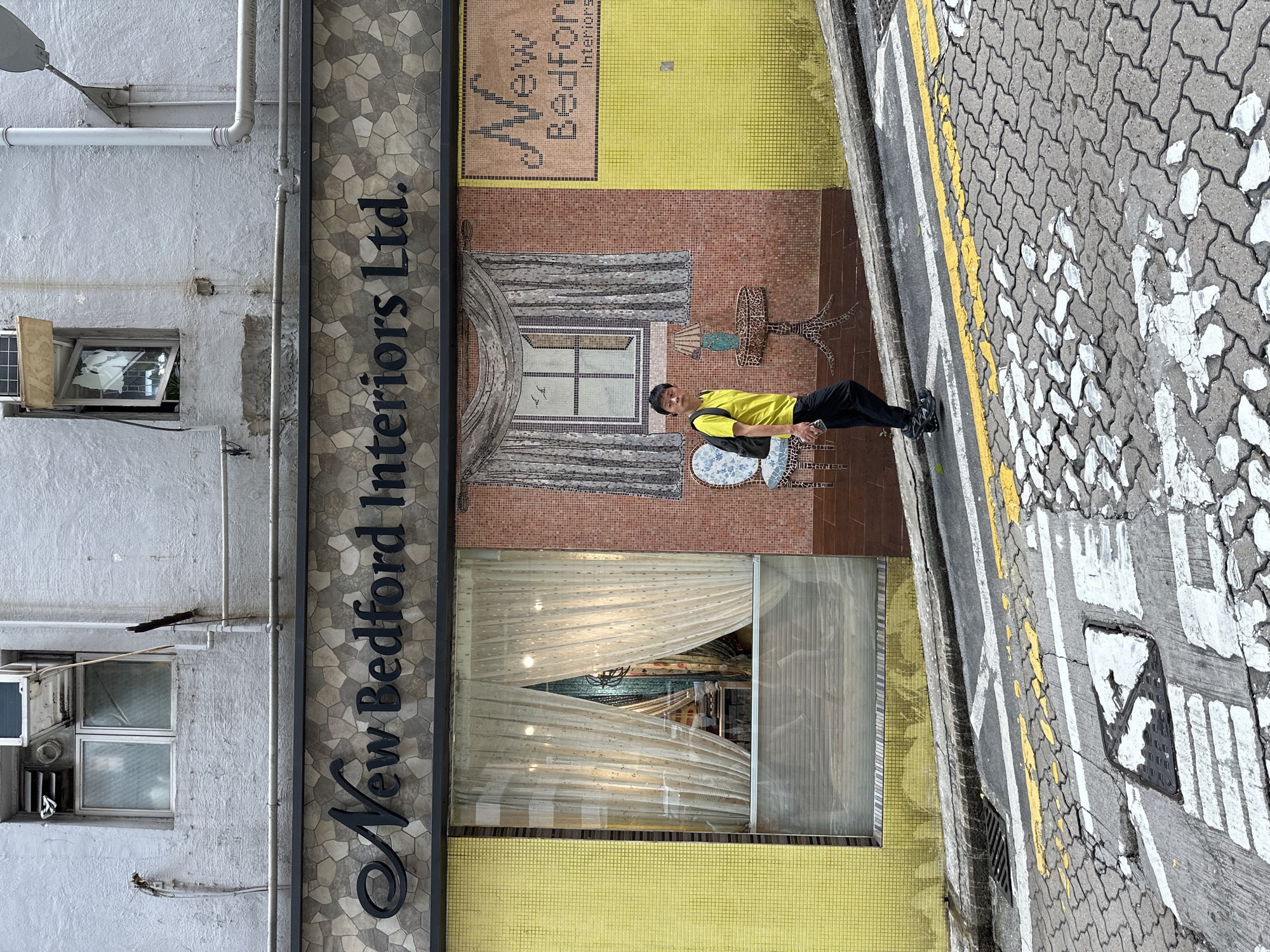 A man wearing a yellow t-shirt and black pants walks on a steep street in Hong Kong. The street is paved with cobblestones and marked with yellow and white lines. The man carries a black backpack and a smartphone. He is walking past a store called "New Bedford Interiors Ltd." The store features a mosaic mural depicting a window with curtains and birds, a chair, and a lamp. The store has a yellow tiled facade and a large window with sheer curtains. The building is old and shows signs of wear, with peeling paint and exposed pipes. The overall scene captures a glimpse of street life in Hong Kong, characterized by its unique architecture and vibrant urban environment.
