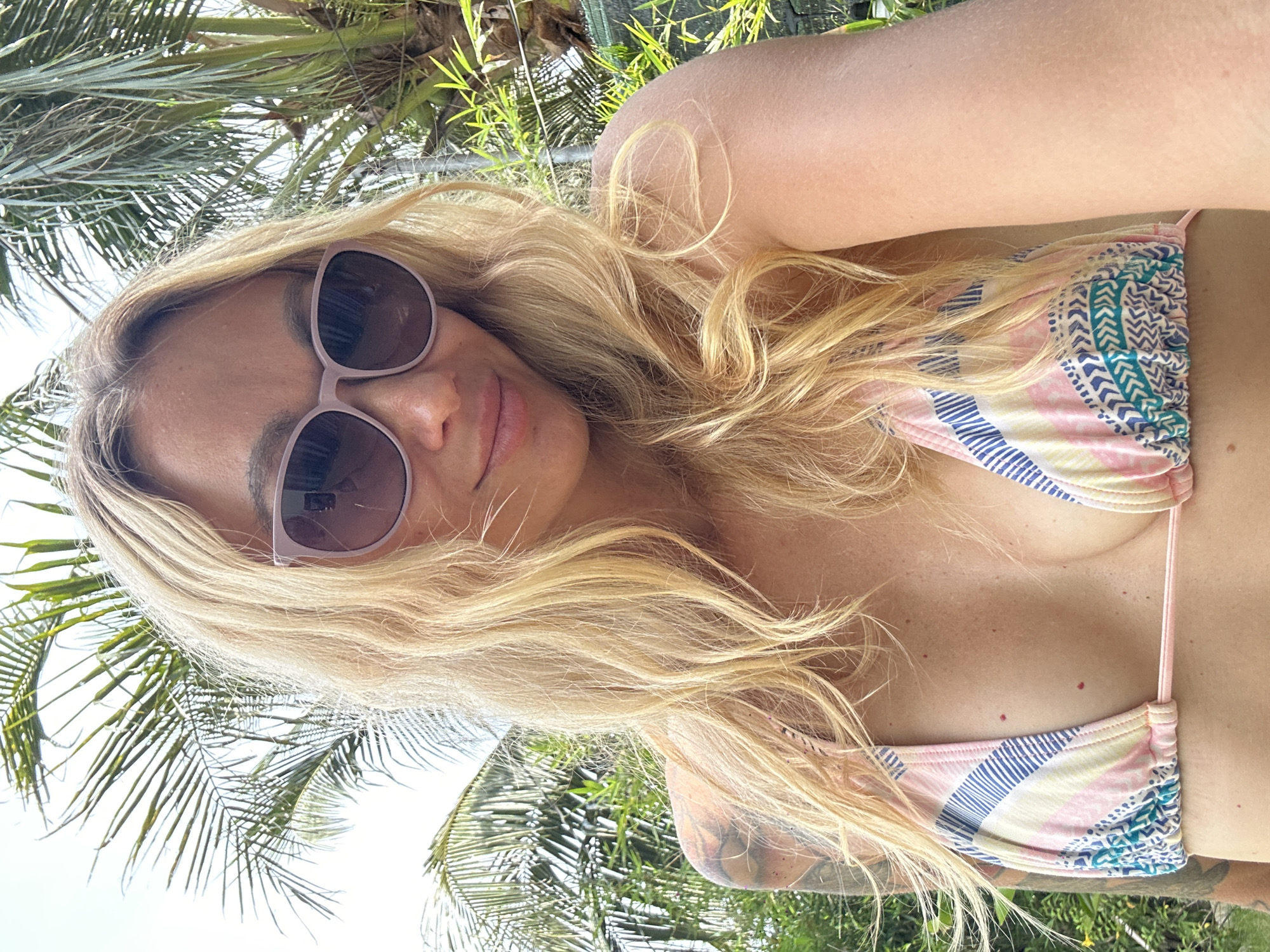 A young blonde woman wearing sunglasses and a pink, blue, and white patterned bikini top poses for a selfie outdoors, with palm trees and lush greenery in the background. The image appears to be taken in Mũi Né, Vietnam, given the tropical foliage.