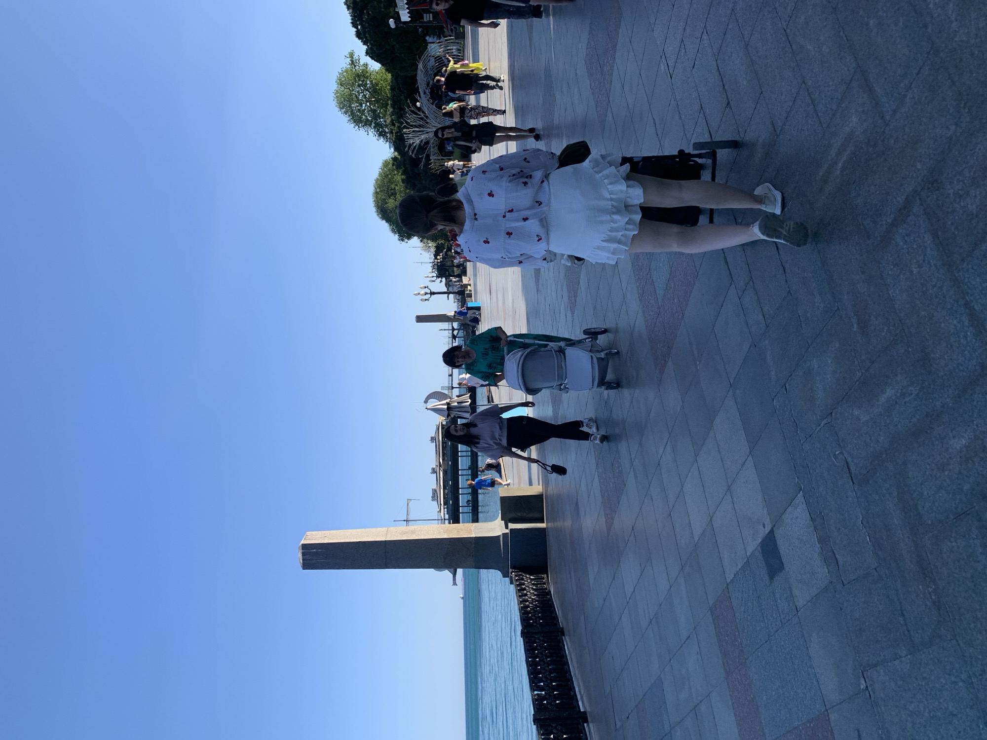 Here is a description of the image:
The image shows a waterfront walkway in Unknown, Unknown. The walkway is paved with gray and red bricks. A stone monument stands on the left side of the walkway. To the left of the walkway is water, and above the walkway is a clear blue sky. The image is shot from a low angle and is oriented vertically.
Several people are walking along the walkway. From the perspective of the shot, they are moving from right to left. Closest to the camera, a woman in a white dress with a cherry print is pushing a black stroller. Further ahead of her, another person is pushing a light blue stroller. Several other people are seen walking on the walkway. Behind the people are trees.