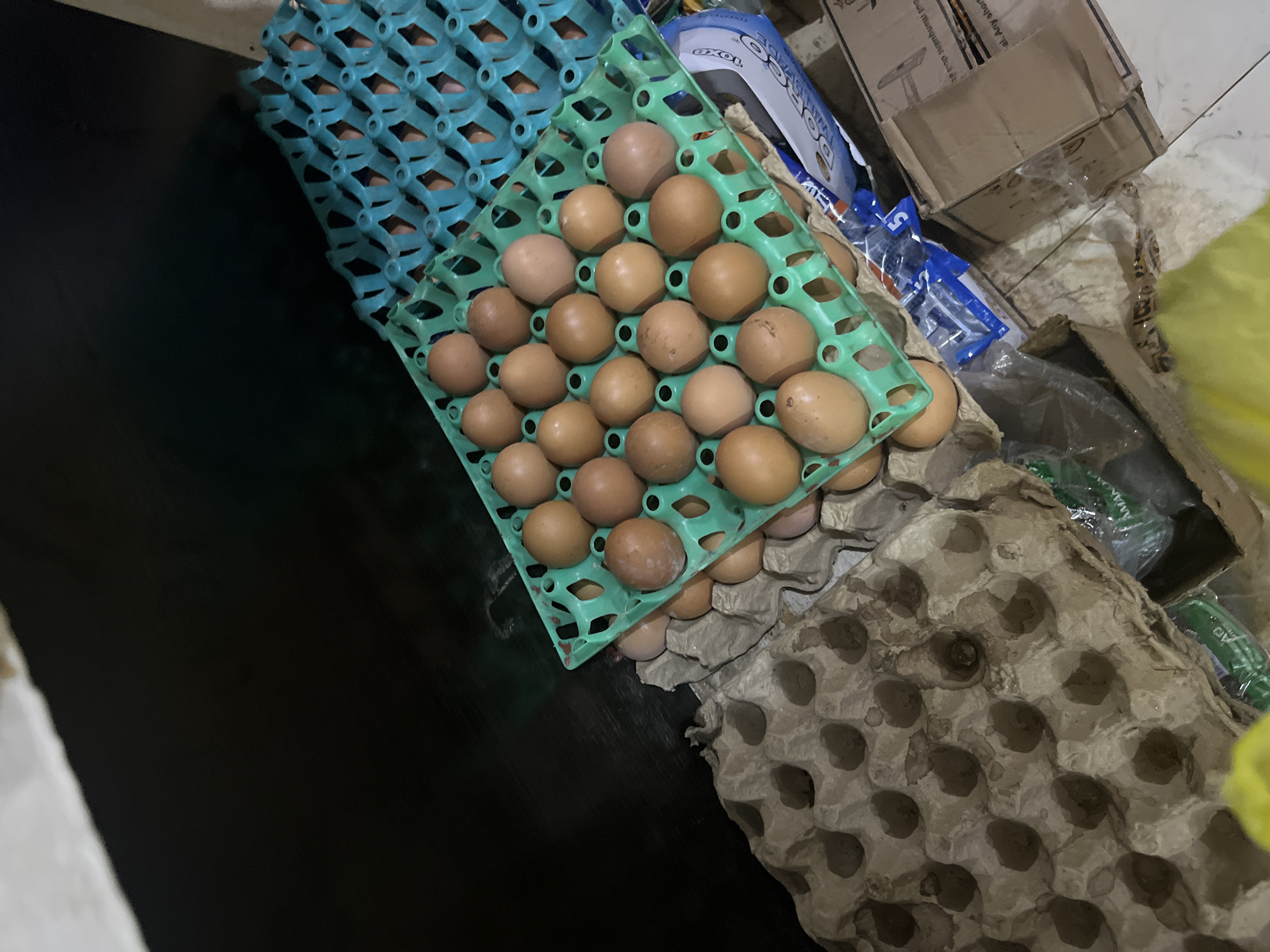 The image depicts a close-up, overhead view of several egg cartons and plastic egg trays filled with brown eggs. The scene appears to be indoors, possibly a market or storage area, with a dark surface in the background and a tiled floor in the foreground. Some of the egg cartons are made of cardboard, while others are plastic and colored green and blue. Several plastic bags and cardboard boxes are also visible, suggesting a commercial setting. The overall lighting is dim, casting shadows on the eggs and cartons. The location is identified as Bukuru, Nigeria. There are no people visible in the image.