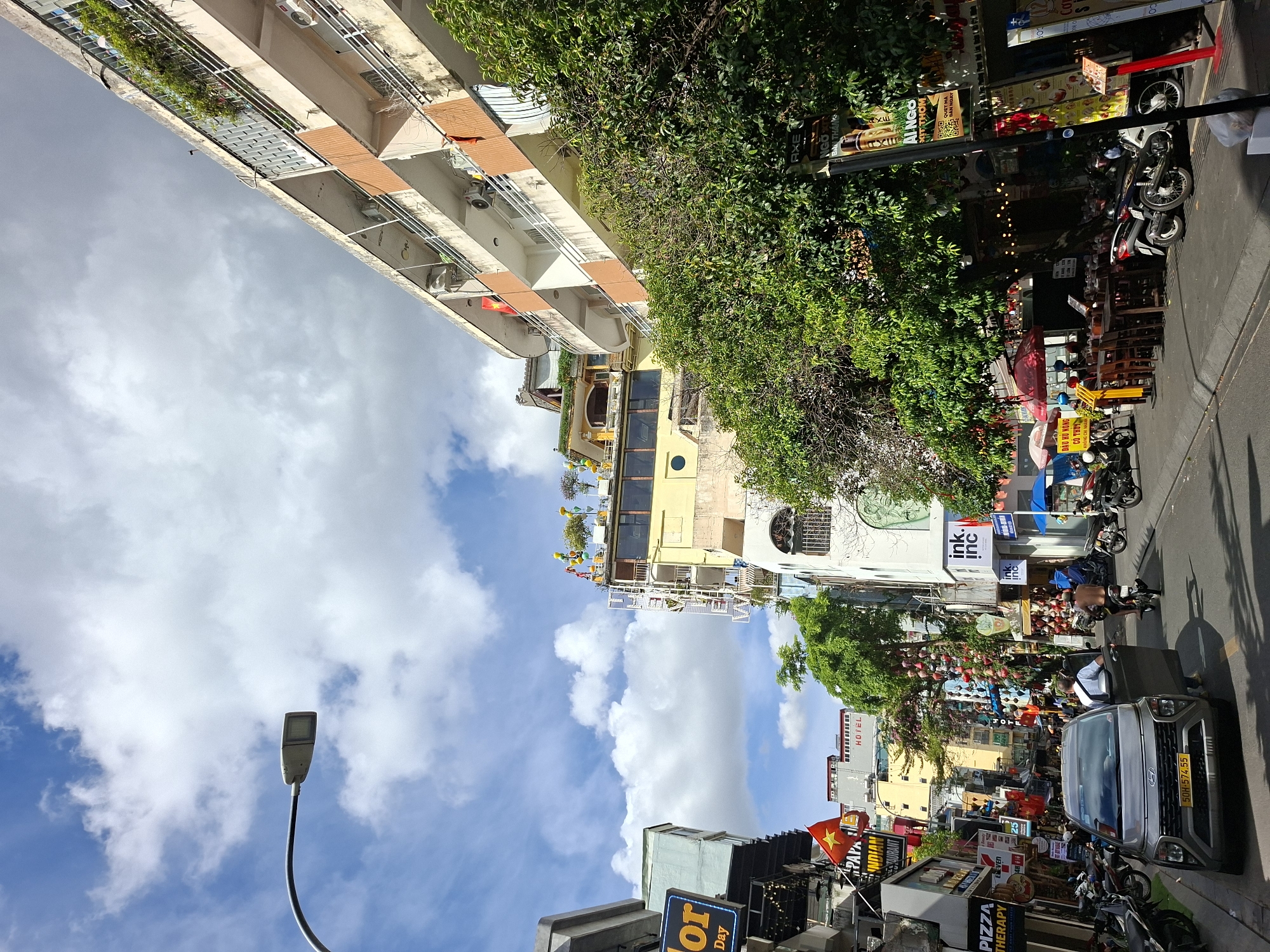 This is a landscape, vertically oriented photograph captured in a bustling street scene, likely in Thủ Đức, Vietnam, during the daytime. The sky is a vibrant blue, dotted with fluffy white clouds, suggesting fair weather.
The foreground is dominated by a street with parked motorcycles and a silver car, its door open, with a person visible inside. A street lamp stands tall, its modern design contrasting with the surrounding environment.
The middle ground presents a vibrant streetscape lined with buildings adorned with various signs and advertisements, indicative of shops and businesses. One prominent sign reads "PIZZA THERAPY ELEVEN," and another displays "HOTEL." The buildings showcase a mix of architectural styles, with some featuring balconies and greenery. Lush trees with red and pink hanging decorations line parts of the street, adding a festive touch.
In the background, more buildings and a glimpse of a taller structure can be seen, further emphasizing the urban setting. The overall impression is one of a lively commercial area, with the bright daylight illuminating the scene and casting shadows. The presence of multiple signs with Vietnamese text, such as "ĐẬU HỦ NÔNG CO THUY" and "AXE NGO BAN THẬP," confirms the Southeast Asian location. The scene appears to be active and full of the energy typical of a city street.