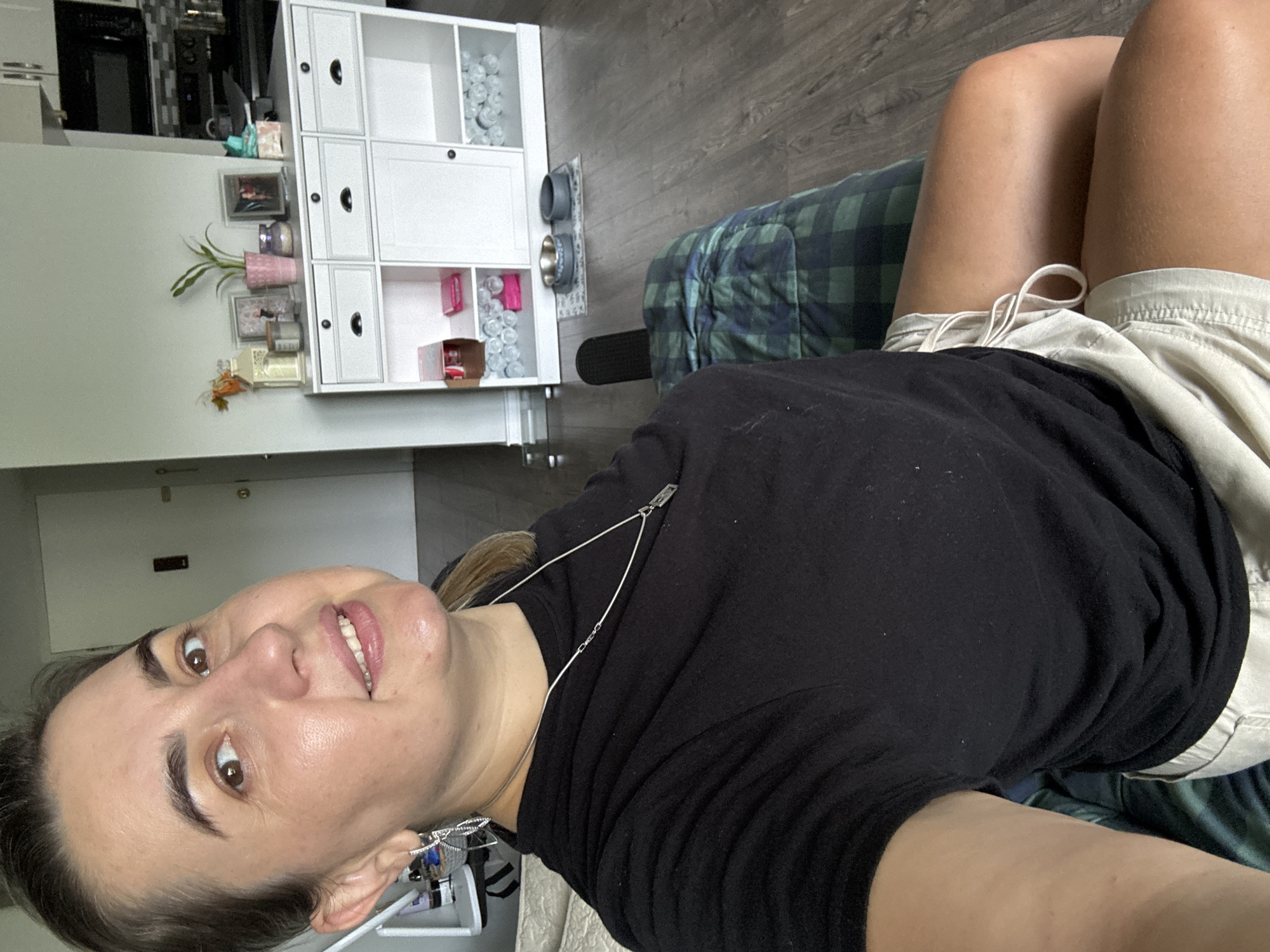 A woman with brown hair and brown eyes is pictured indoors, likely in a living room or bedroom. She is wearing a black t-shirt and light-colored shorts. Her left arm is resting on a plaid-patterned surface. In the background, there is a white cabinet with shelves and drawers. The floor appears to be wood laminate. The image is taken from a slightly high angle, looking down at the woman. There is no indication of the specific location in the city of Wheeling, United States of America.