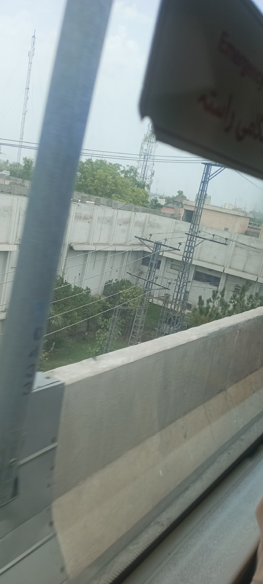 The image shows a view from inside a vehicle, looking outwards. In the foreground, a gray metallic structure, possibly part of the vehicle's interior or an external railing, is visible on the left. A blurred concrete barrier runs horizontally across the lower portion of the image. Beyond the barrier, a building with a light-colored facade and a flat roof is seen. Several large electrical pylons or transmission towers are scattered throughout the scene. Green trees and vegetation are present between the structures and in the background. The sky is overcast. A small portion of a red and white sign is visible in the upper right corner, with text that appears to be in a foreign language. The overall impression is an urban or semi-urban environment, possibly in Alam Khan Kalay, Pakistan, based on the context provided.