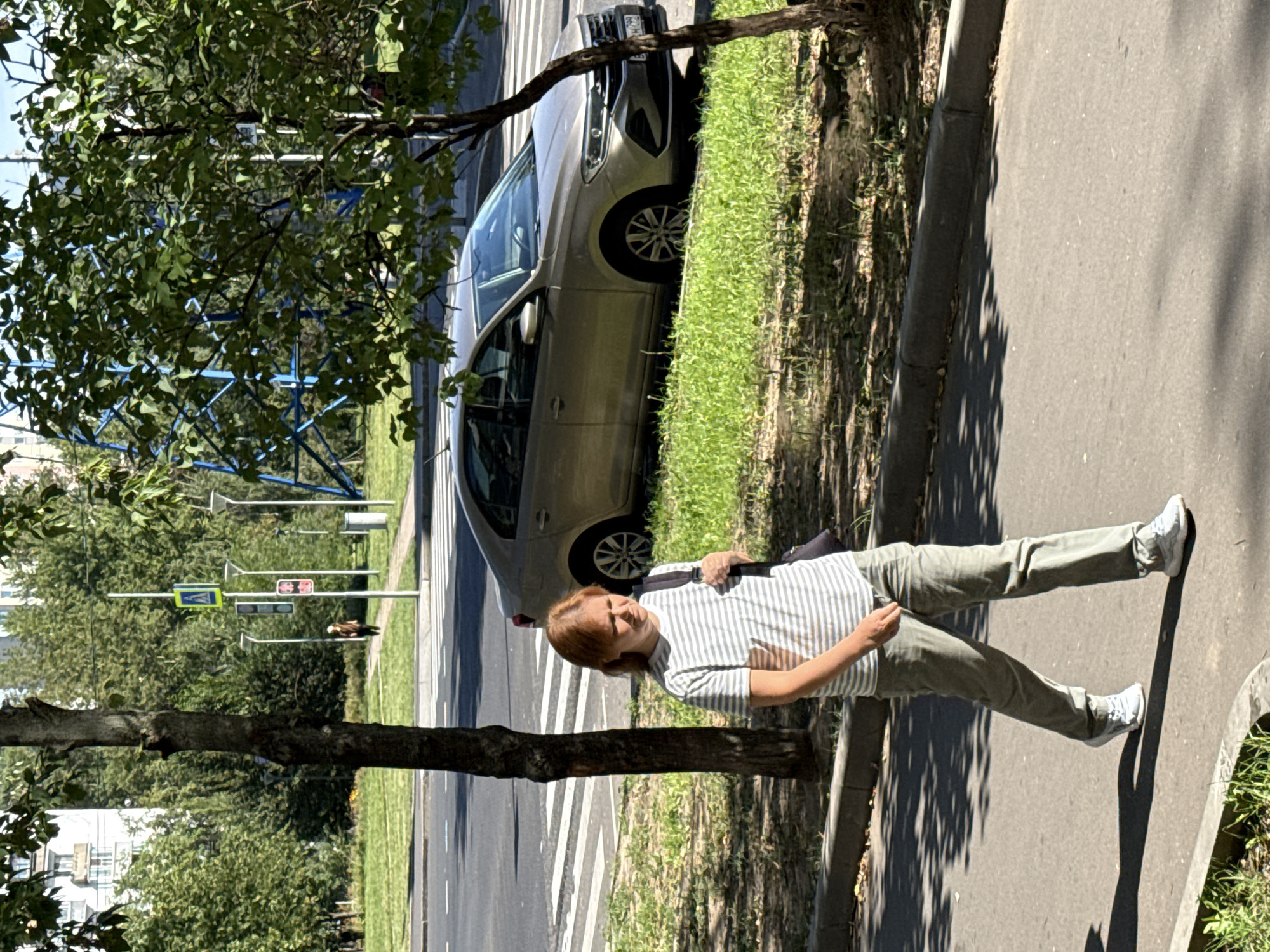 A woman with reddish-brown hair is walking on a sidewalk in Moscow, Russia. She is wearing a striped t-shirt and light-colored pants. She is carrying a dark bag over her shoulder. She is walking on a sidewalk next to a street with a pedestrian crossing. A light-colored sedan is parked on the side of the street. There are trees and grass around the sidewalk and street. In the background, there are buildings and street lights. The sun is casting shadows on the ground.