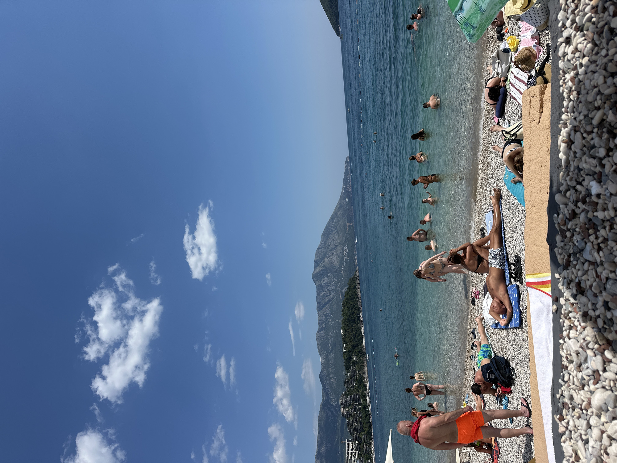 This is a wide-angle, landscape-oriented photograph taken from a beach in Budva, Montenegro. The image is vertically oriented within the frame. A clear blue sky with scattered white clouds occupies the left portion of the image. The right side features a pebbled beach where numerous people are relaxing and swimming in the turquoise, clear water of the Adriatic Sea.
On the beach, individuals are sunbathing on towels and enjoying the seaside. Some are in the shallow water, wading or swimming. In the background, a long, mountainous coastline stretches across the horizon, dotted with lush green trees and buildings, indicative of a coastal town. The sea is calm, with gentle waves lapping the shore. The sunlight suggests it is a warm, sunny day, likely mid-morning to early afternoon. The atmosphere appears relaxed and leisurely, with people enjoying a typical summer day at the beach.
There are no visible text elements in the image. The focus is on the natural beauty of the location and the recreational activities of the beachgoers.