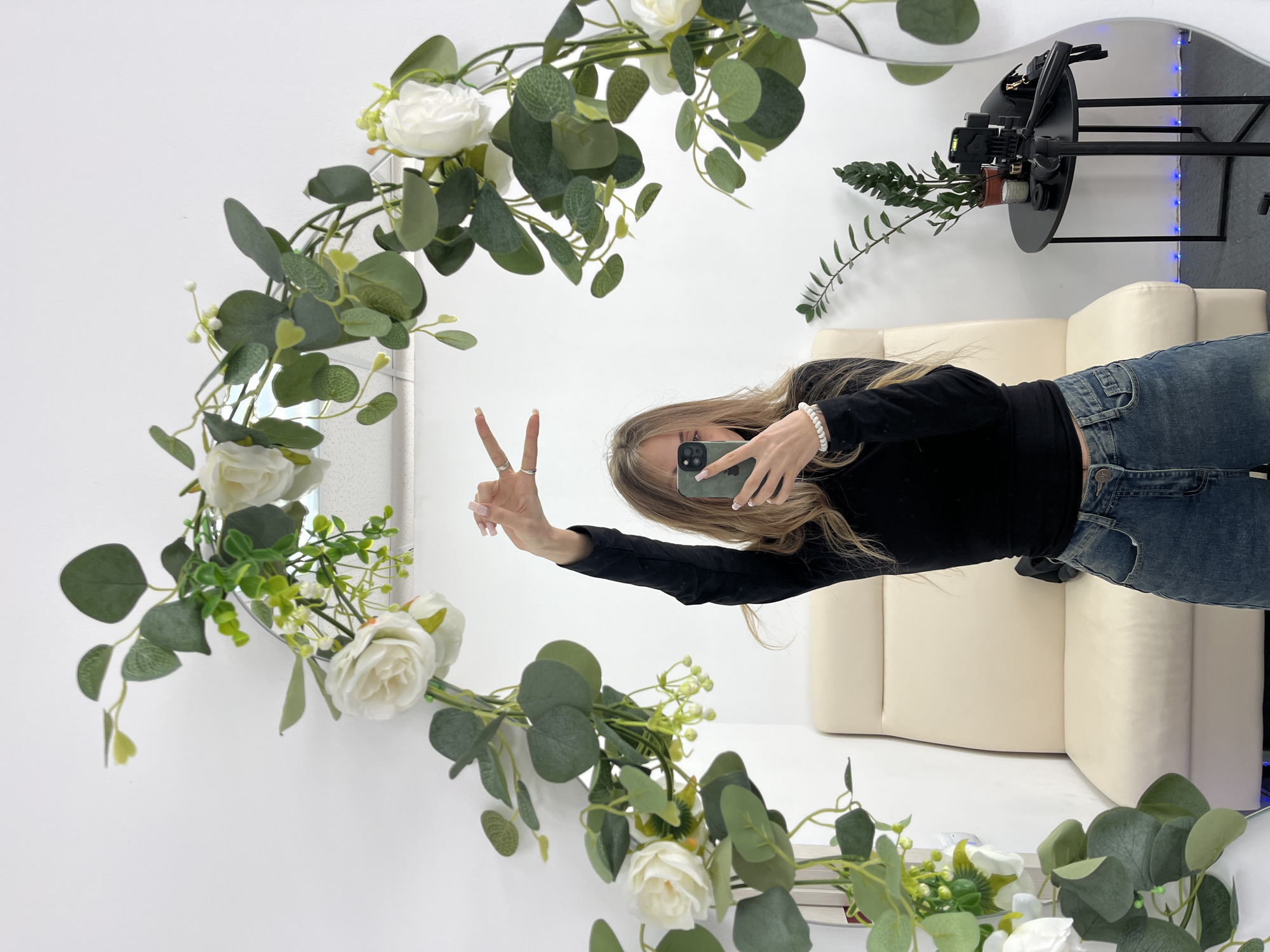 A female subject is captured in a mirror selfie within an indoor setting in Фоки, Russia. She stands centrally, holding a green-cased smartphone in her right hand to photograph her reflection. Her left hand is raised, fingers forming a "peace" sign, adorned with two rings. She has long, light brown hair and is dressed in a black long-sleeve top, faded blue jeans, and a white beaded bracelet on her right wrist.
The room features white walls and a dark grey floor, with a subtle blue LED light strip visible at the base of the wall on the right. The large mirror, which reflects the subject, is framed along its top and sides by an artificial garland composed of white roses, green eucalyptus leaves, and small light green berry-like clusters. To the subject's left, a high-backed, upholstered beige chair or treatment bed is present. To her right, a small black oval side table holds a potted green plant, a black camera tripod with a phone holder, and various small dark objects, potentially camera accessories or cosmetic items.