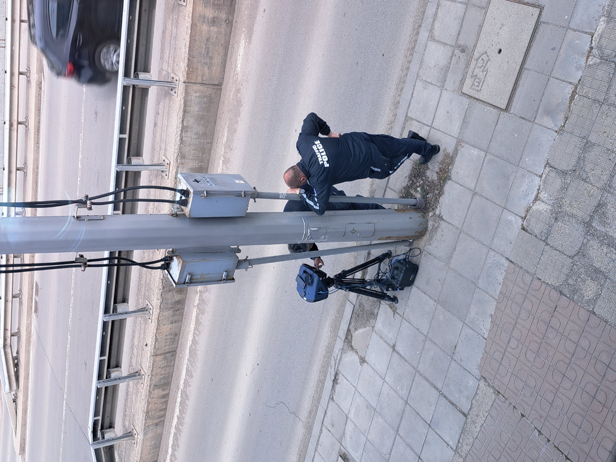 This overhead image, taken in Sofia, Bulgaria, depicts a traffic police officer engaged in speed enforcement activities. A male officer, wearing a dark blue uniform with "TRAFFIC POLICE" emblazoned in white across his back, is positioned beside a concrete road, leaning against a tall, gray utility pole. He appears to be looking downwards, likely attending to his equipment or observing traffic.
Adjacent to the officer, on a paved sidewalk, is a portable speed monitoring setup. This consists of a black tripod supporting a blue-cased device, presumably a speed camera or radar gun, which is connected by wires to a black power unit or battery pack resting on the ground. Two gray electrical boxes are also visible, mounted on the utility pole, one with a faint '+' symbol and partially obscured text on its surface.
In the blurred background, a dark-colored vehicle is seen moving on the road, alongside concrete barriers and metal guardrails, indicating a road, possibly an overpass or highway. The sidewalk features a mix of gray and patterned brown paving tiles. The scene occurs during daytime under clear weather conditions, highlighting an active traffic surveillance operation.
