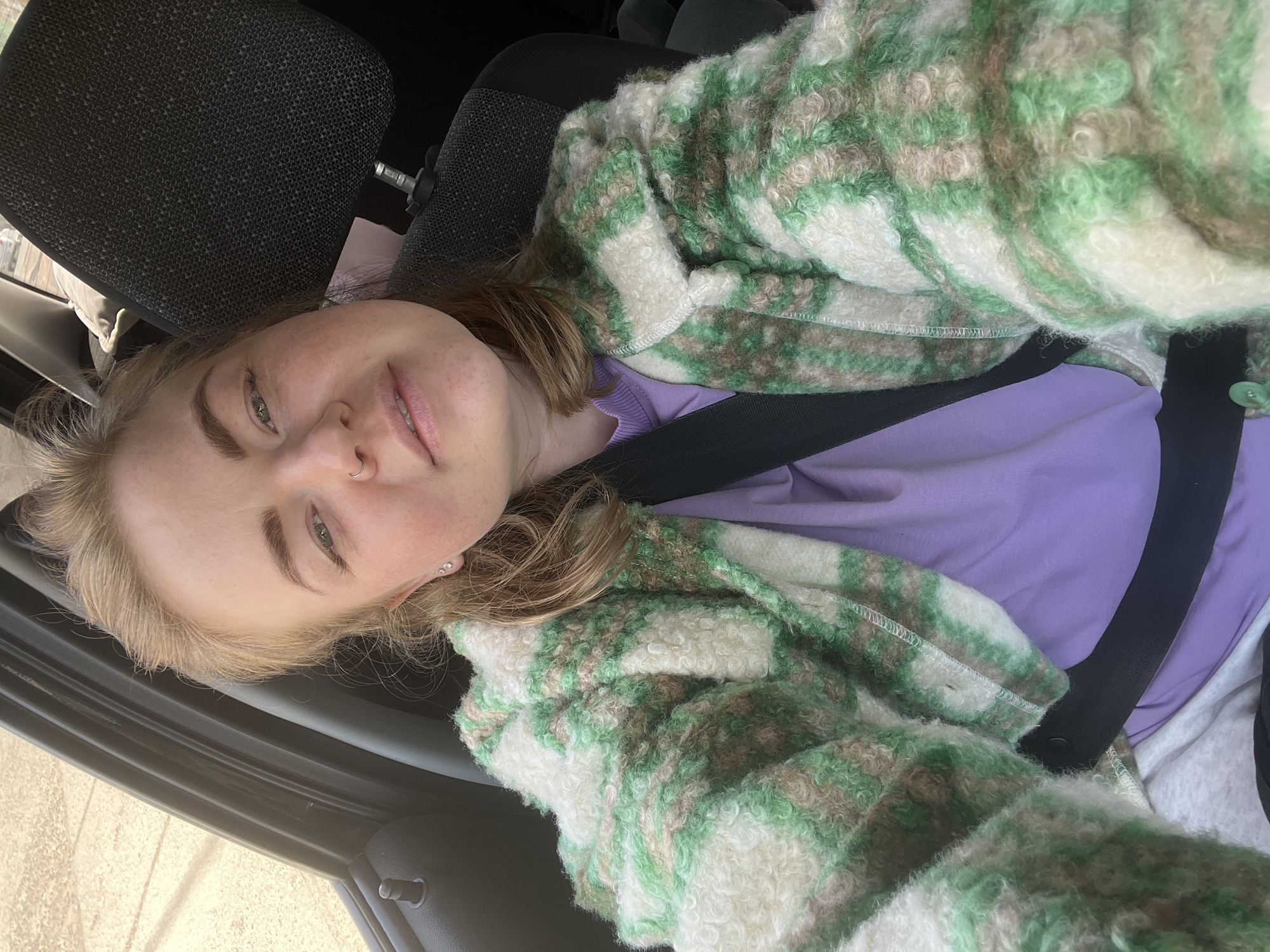 A young Caucasian woman wearing a purple t-shirt and a green and white striped jacket is in a car, buckled in with a black seatbelt. She is looking at the camera, smiling slightly. The interior of the car includes a grey seat headrest. The location is not evident and the city cannot be determined.