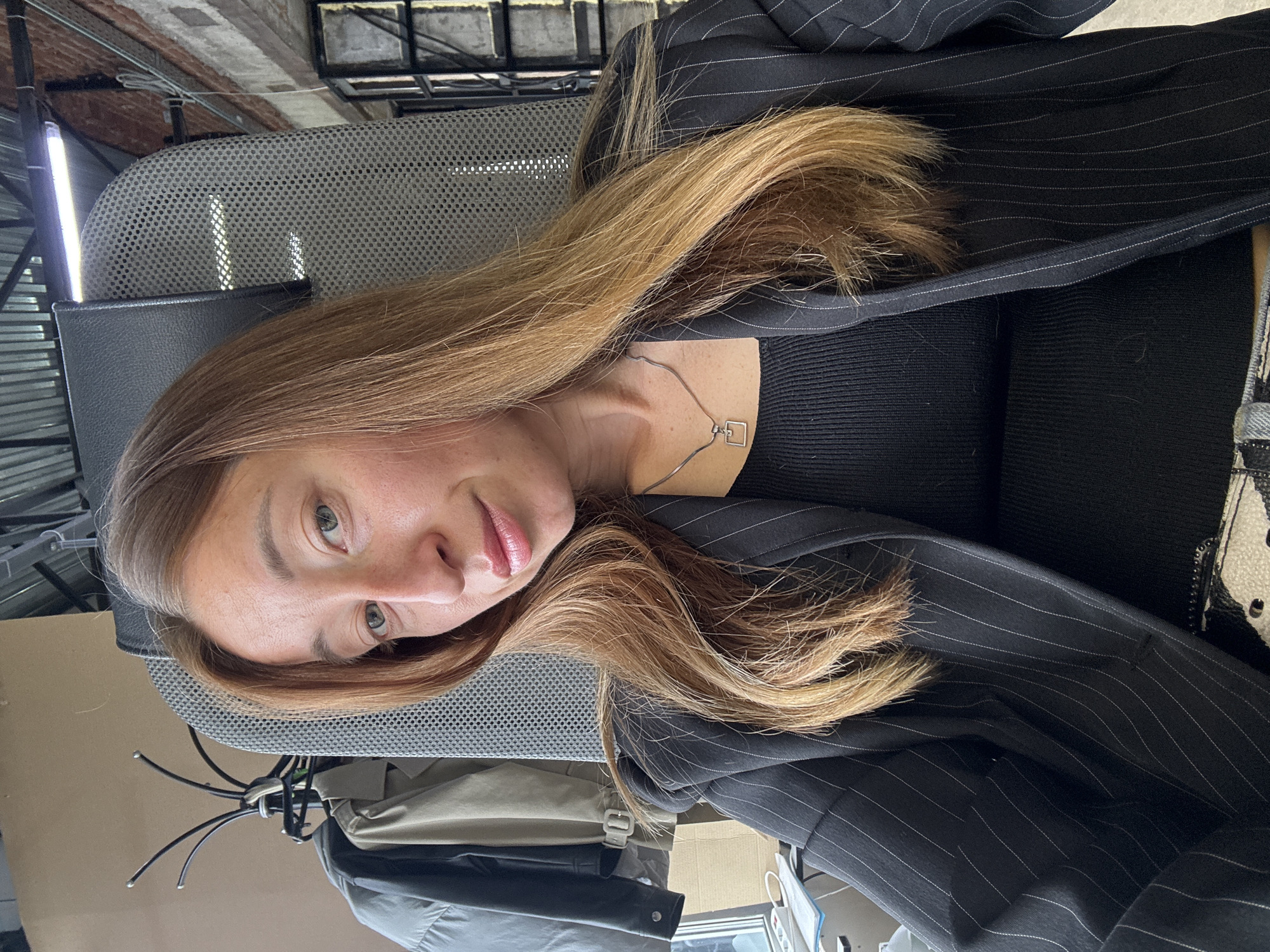 A woman is captured in a selfie, likely indoors. She has light brown hair and fair skin. She is wearing a black ribbed top and a black pinstripe blazer. A silver necklace is visible. The setting seems to be an office environment, with a grey mesh office chair and other articles of clothing visible in the periphery. It is impossible to determine if the location is in the city of Chaikovsky, Russia, from this image.