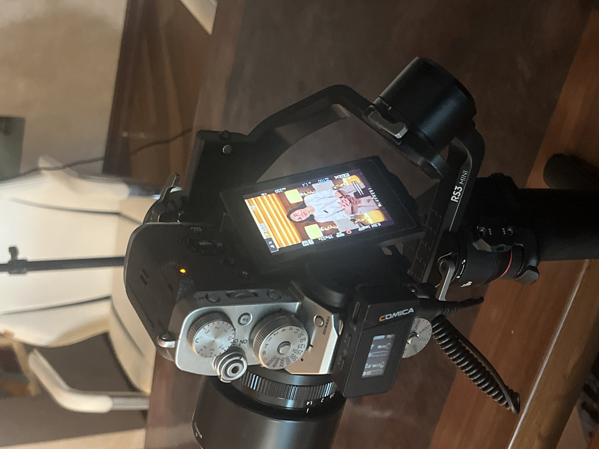 The image shows a close-up of a camera setup on a wooden table. A camera with a large lens is mounted on a gimbal stabilizer. The camera's screen displays a woman in a white shirt, likely being filmed. The background is blurred, showing what appears to be an indoor setting with furniture. There is no discernible information in the image to suggest a location in the city of Kazan, Russia.