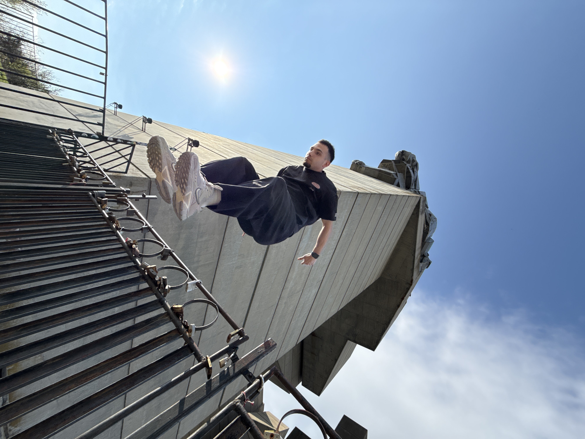 The photograph, taken from a low angle, depicts a man suspended in mid-air against the backdrop of the Monument to 1300 Years of Bulgaria in Shumen, Bulgaria. The monument, a large concrete structure with angular and abstract sculptural elements, dominates the right side of the frame. The sky, a clear blue with scattered clouds and the sun shining brightly, fills the background. To the left, a metal structure, possibly part of the monument's access or maintenance system, extends upwards, revealing a grid-like pattern. The man, dressed in dark clothing and light-colored sneakers, is oriented horizontally, his body seemingly parallel to the monument's surface. He is positioned near the top of the metal structure.