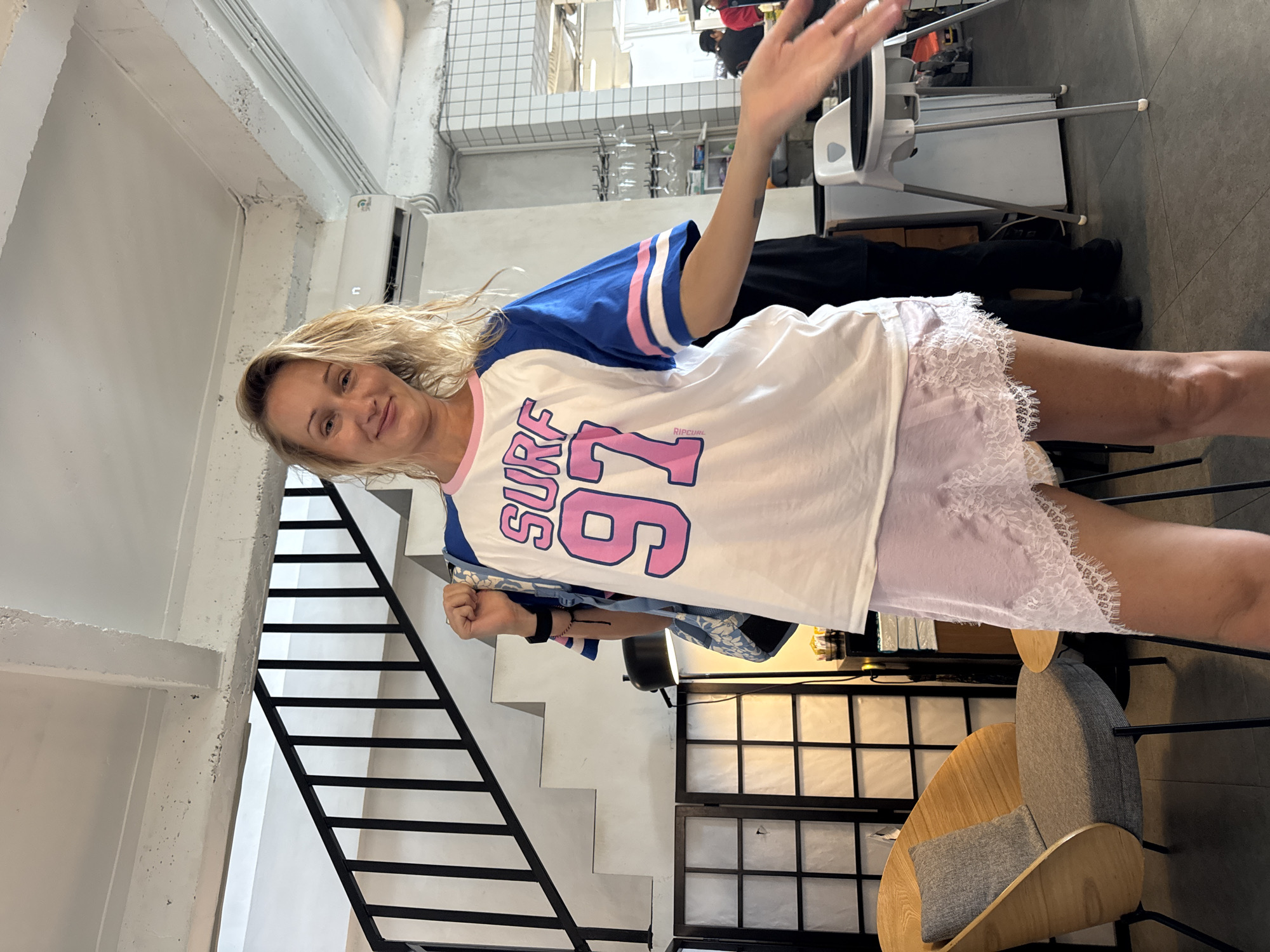A blonde woman is posing indoors, holding her hand up in a greeting gesture. She is wearing a white t-shirt with blue sleeves and pink lettering and appears to be wearing shorts with lace trim. In the background are stairs, a decorative screen, and a chair. The location is Kuta Selatan, Indonesia.