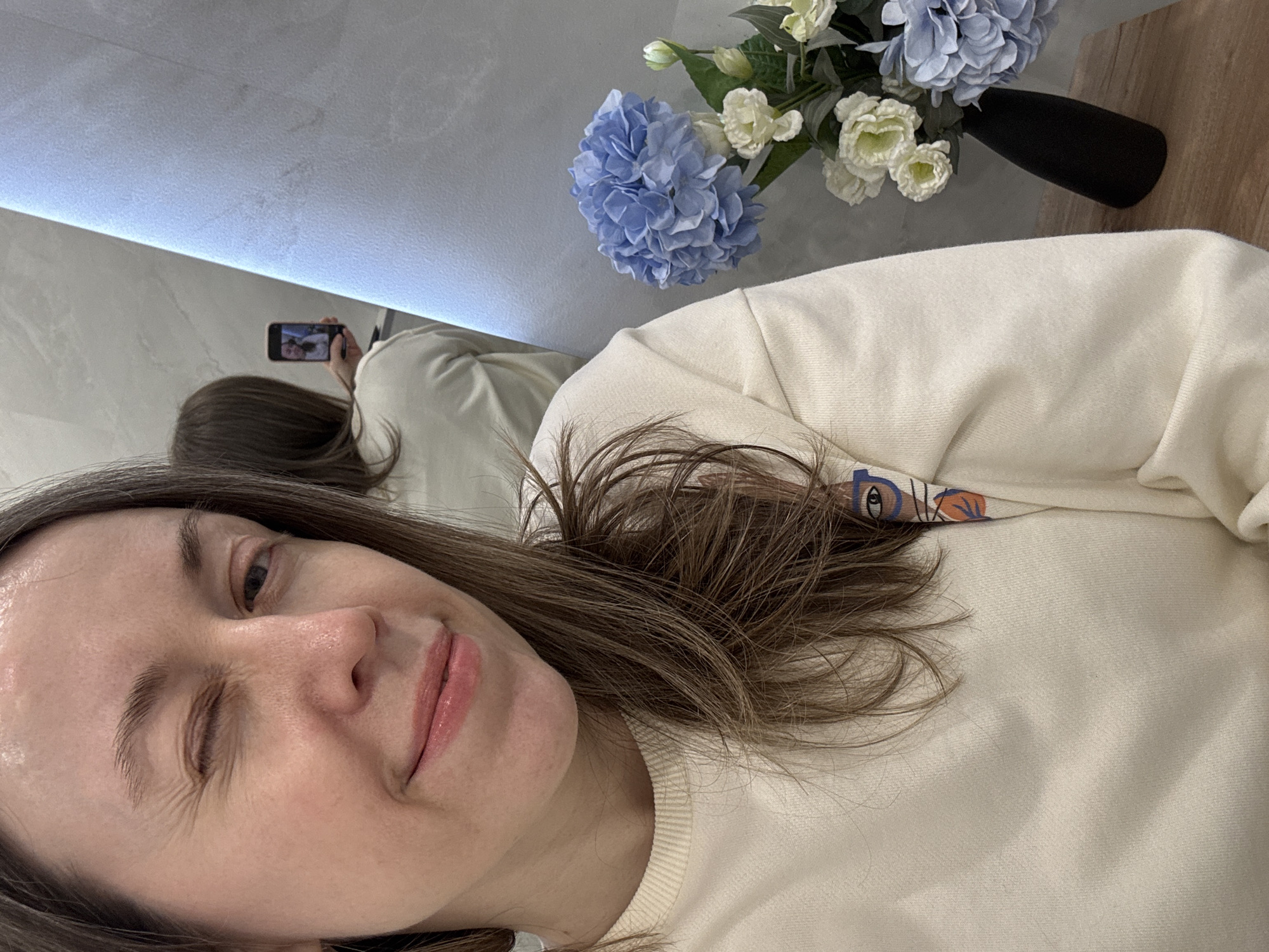 A selfie of a woman with brown hair, winking, wearing a cream-colored sweater, with a bouquet of blue and white flowers in a black vase visible in the background. The woman is also reflected in a mirror in the background, where she appears to be holding a phone taking the selfie. The location, Saint Petersburg, Russia, cannot be determined from the visual information provided.