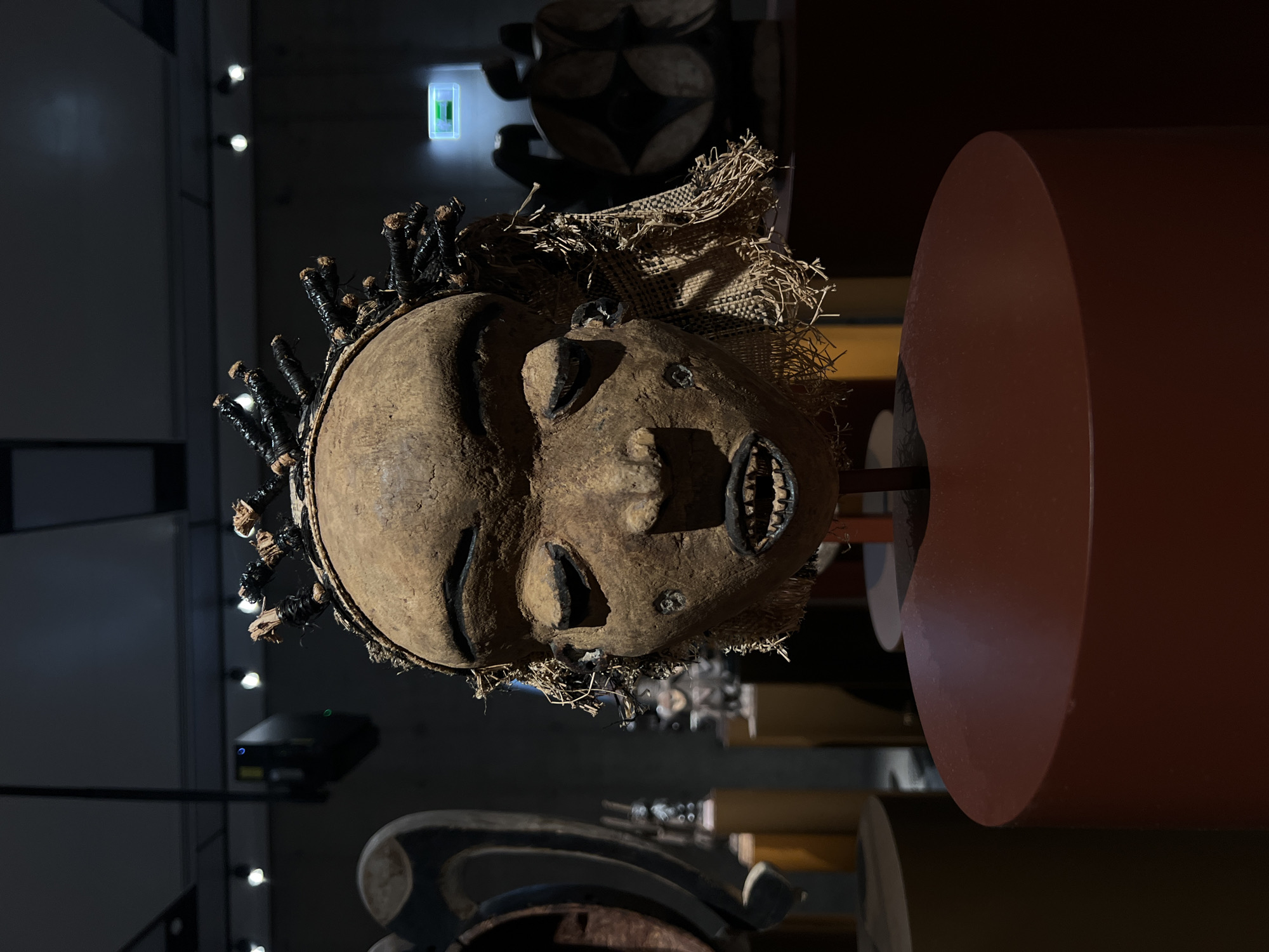 A vertical, low-angle shot depicts an artifact, an aged tribal mask, within a museum setting in an unknown city. The mask, predominantly brown in tone, features coarse, textured skin, carved facial features, and decorative elements resembling hair bound with dark wrappings. A portion of burlap-like material adorns the upper section of the mask. The mask is mounted on a thin, dark metal rod that is attached to a reddish-brown, cylindrical display stand. The museum environment is dimly lit. In the background, additional cylindrical display stands, along with other tribal artifacts, are vaguely visible. A black projector hangs from the ceiling on the left side of the frame. A rectangular illuminated sign is visible high on the wall, behind the mask. The overall composition suggests a cultural exhibit focused on tribal art.