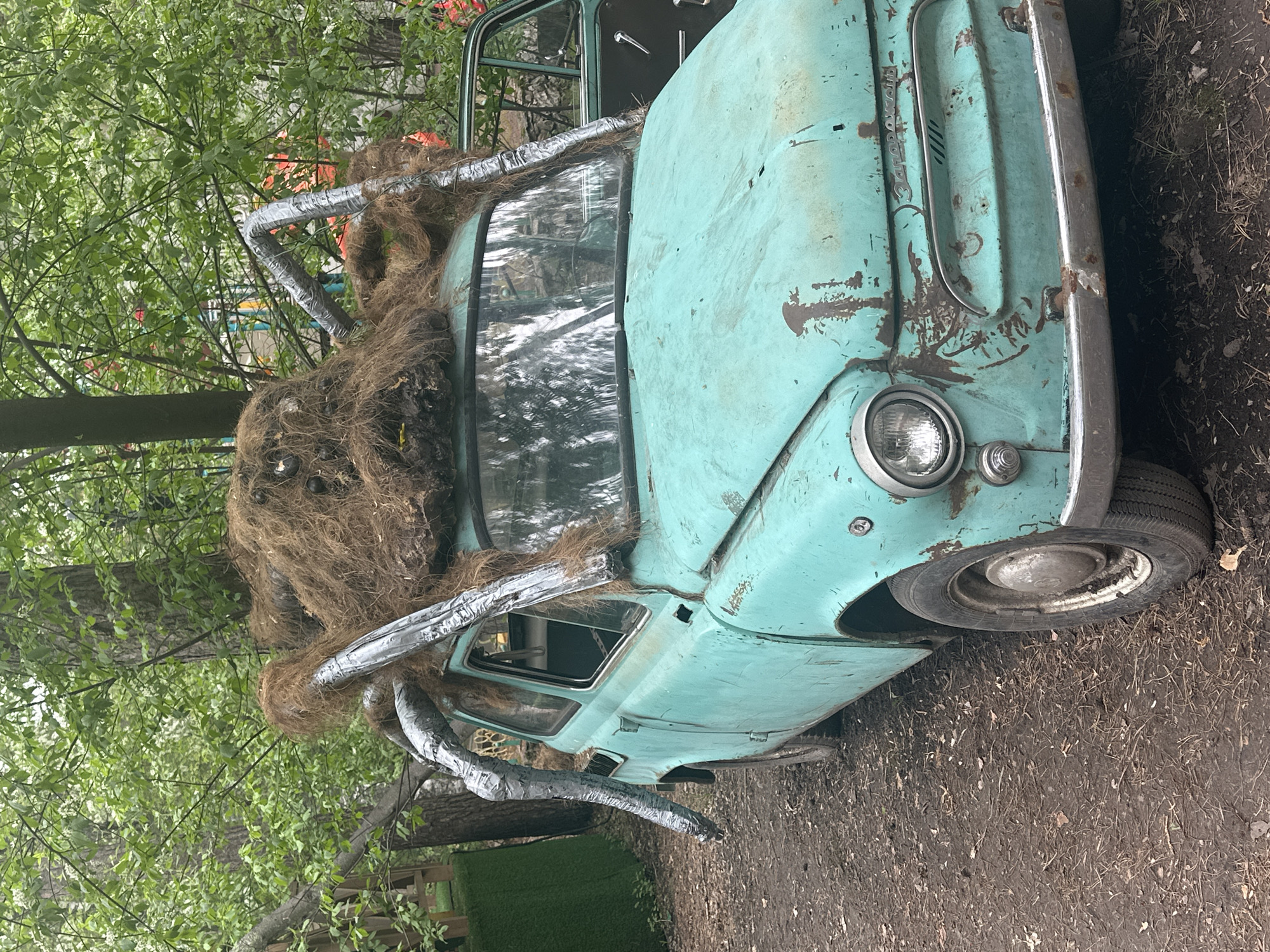 An altered, teal-colored Zaporozhets automobile rests in a dirt and wooded area. The car is decorated with a large, brown, hairy spider-like figure on top, with legs made of silver-duct-taped pipes extending down the windshield and sides. The car appears weathered and slightly rusted. Foliage from surrounding trees frames the car. The location is identified as Berdsk municipality, Russia.