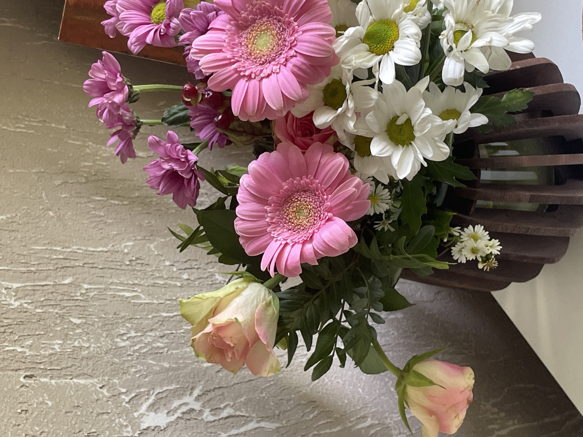 A close-up, eye-level, medium shot captures a bouquet of mixed flowers arranged in a dark brown, wooden vase with a fluted design. The vase is placed on a white surface, against a textured, beige wall with white and gray streaks. The bouquet is predominantly composed of pink gerbera daisies with bright yellow centers, interspersed with smaller white daisies featuring green centers. Several small, pink, daisy-like flowers and some dark red berries are also visible. A single pale pink rose with a yellowish-green center is positioned on the left side of the bouquet, with its stem drooping slightly. Another similar rose, also pale pink, is visible lower down on the left, facing left. Lush green leaves fill the spaces between the flowers. The lighting is soft, casting subtle shadows and highlighting the textures of both the flowers and the wall.