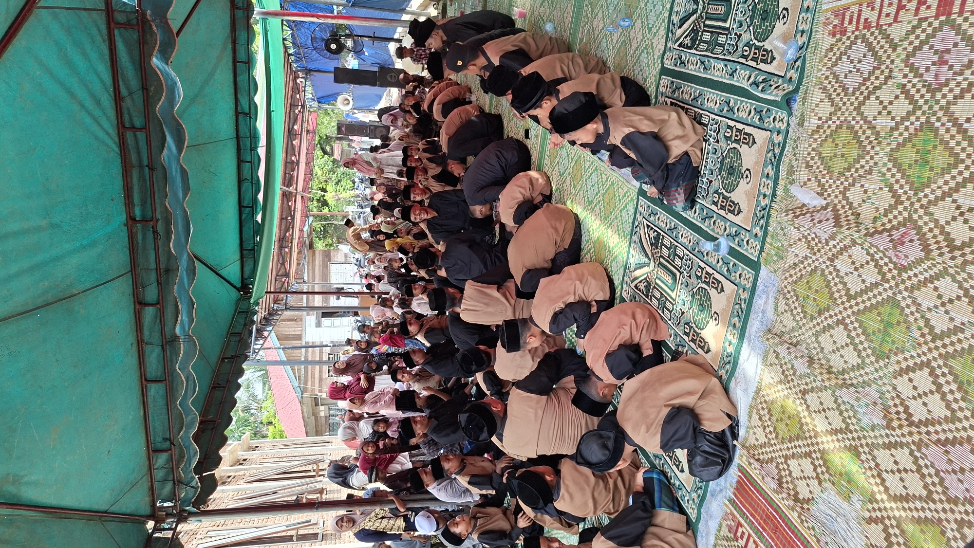The image depicts an outdoor gathering of people, predominantly young males dressed in brown and black attire, sitting on prayer mats under a green tarp. The setting appears to be in Asan, Indonesia, evidenced by the presence of buildings with traditional Indonesian architecture in the background and a predominantly Indonesian population. The individuals are seated in groups, facing different directions, with some engaged in prayer or listening to an unseen speaker. Several adults, including women in hijabs, are standing and observing the gathering from the sides. The environment includes a temporary structure with a tarp providing shade, suggesting an event like a religious ceremony or a community gathering. The ground is covered with various mats, including intricately patterned prayer mats and woven mats. The overall scene conveys a sense of community and religious observance.